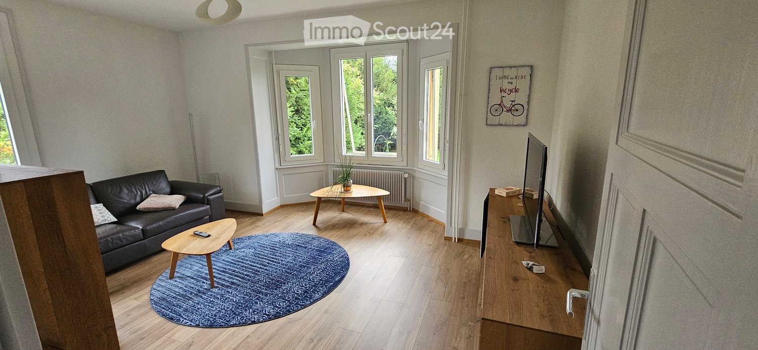 Living room with wooden flooring, large windows, a black leather sofa, a coffee table, a small round table, a screen, and a blue rug.