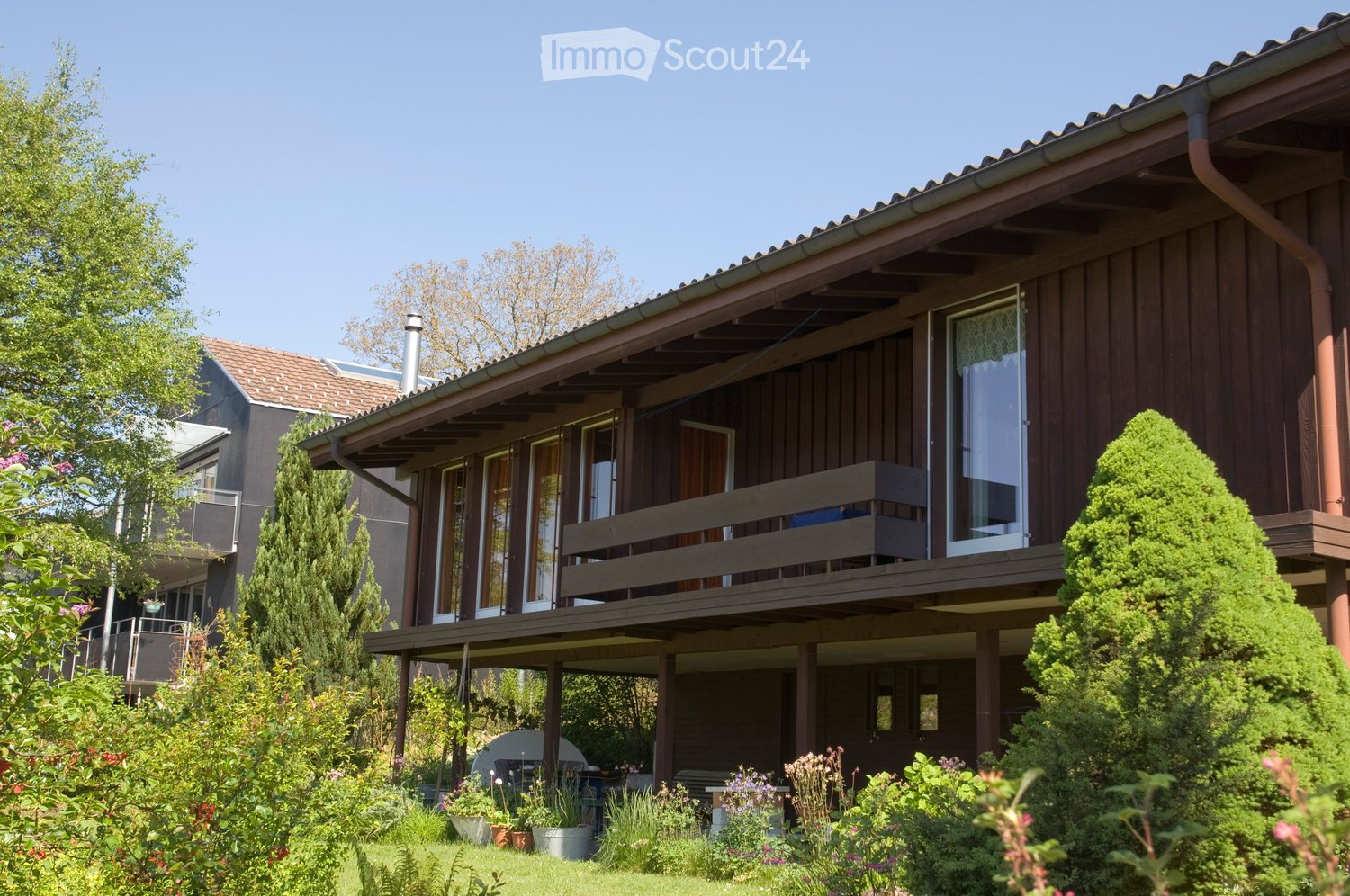 Two-story wooden house with a raised ground floor, large windows, and a covered balcony. The garden features various plants, shrubs, and a small seating area.