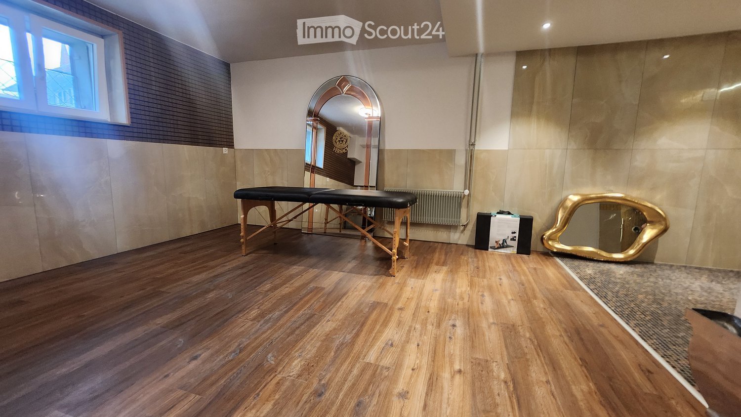 A well-lit room with wooden flooring, a massage table, a large arched mirror, a gold-framed mirror, and a radiator.