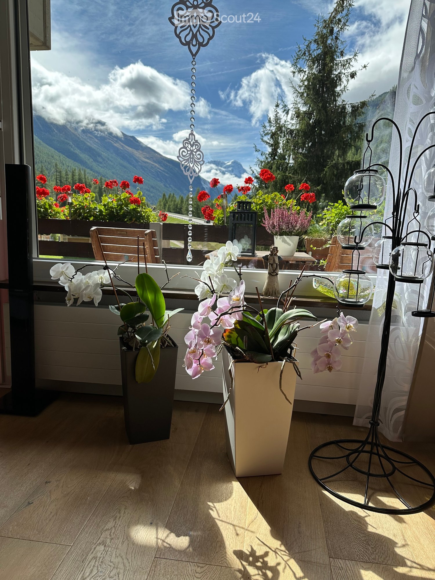 Balcony with wooden floor, large window showing mountain view, decorated with potted orchids, lanterns, and a chair.
