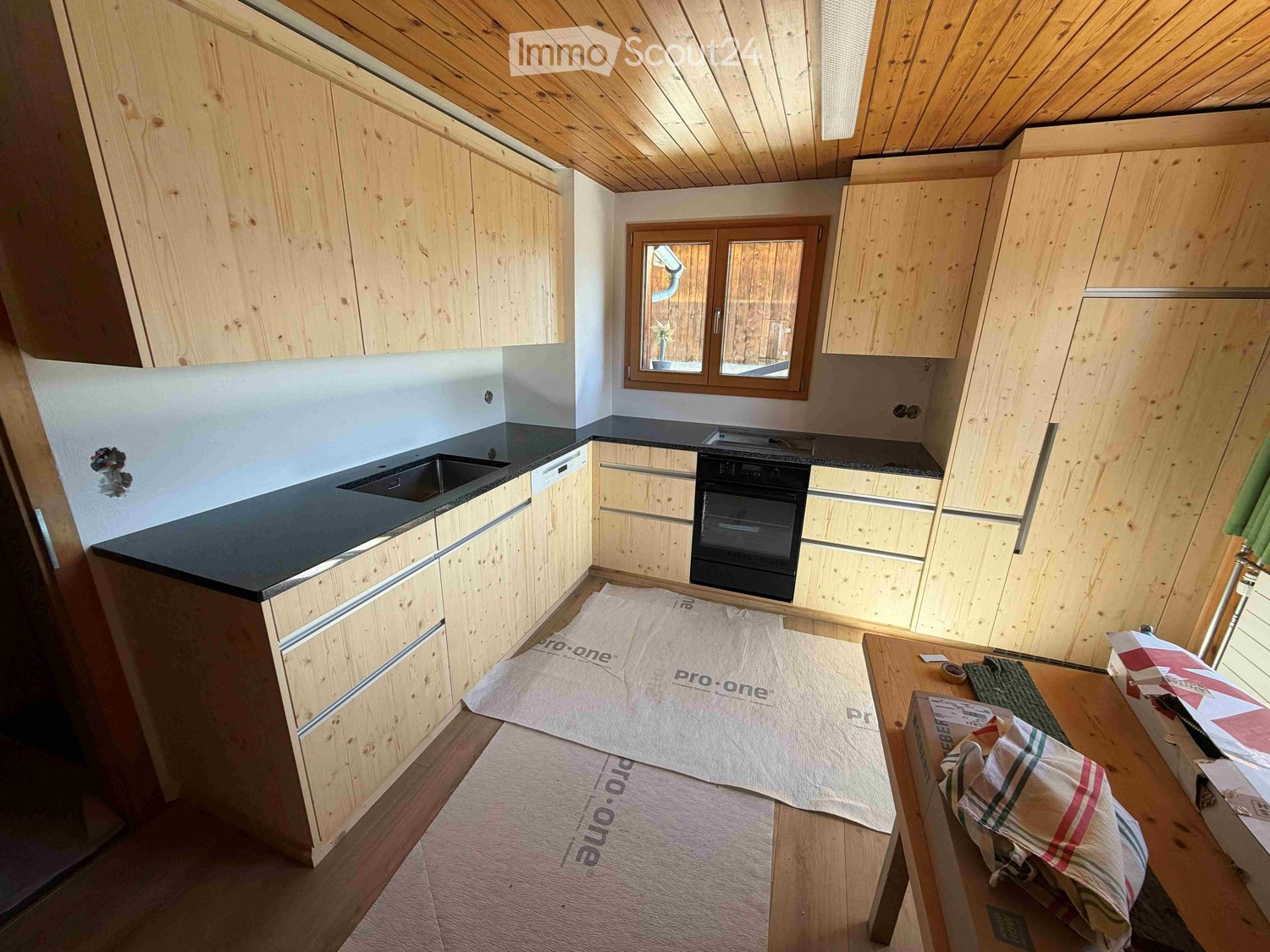 Modern kitchen with wooden cabinets, black countertops, and stainless steel appliances. The space features a sink, oven, and ample cabinet storage. The floor is covered with protective sheets.
