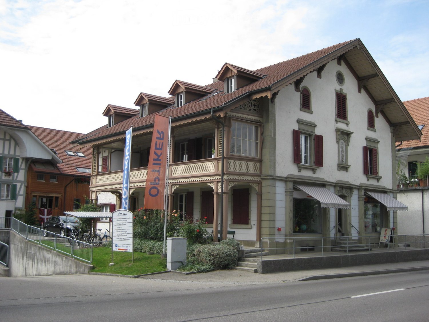 Two story house with brown roof, white walls, red shutters, brown wooden balcony, green lawn, and banners on the front.