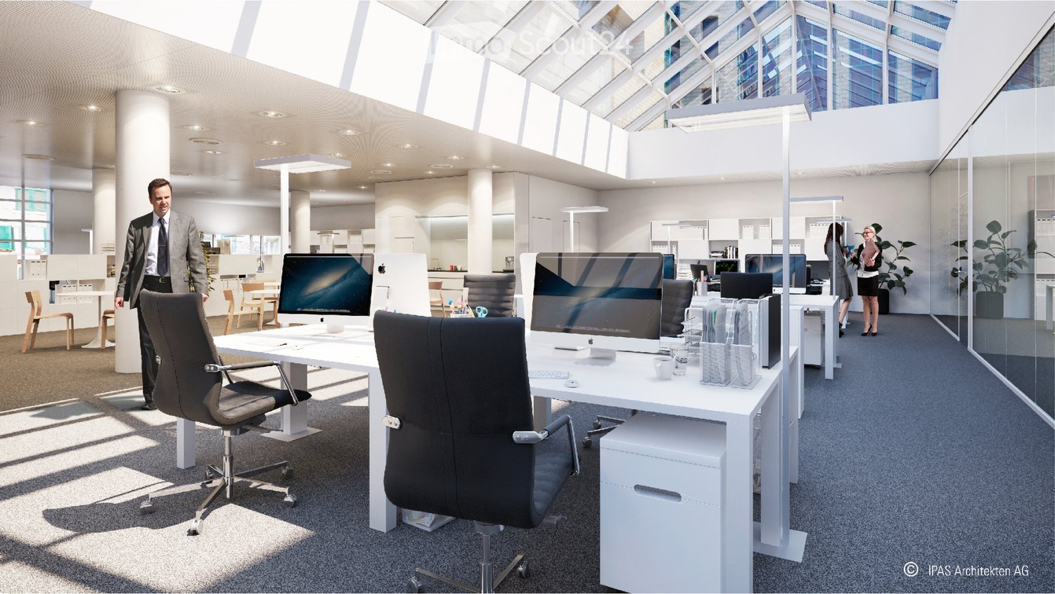 Modern office space with white desks, black office chairs, computer setups, and large windows allowing natural light. There are also potted plants and a kitchen area visible in the background.