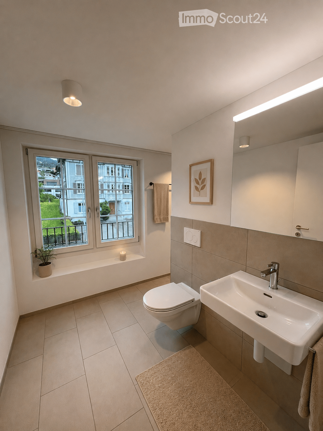 Modern bathroom with a toilet, sink, and a window offering a view of the outside. Includes a towel, a small plant, a picture frame, and a rug on the floor.