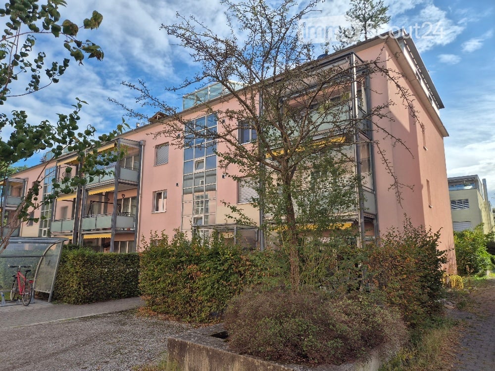 Modern multi-story residential building with pink facade, large windows, and balconies. Surrounded by greenery and a paved pathway.