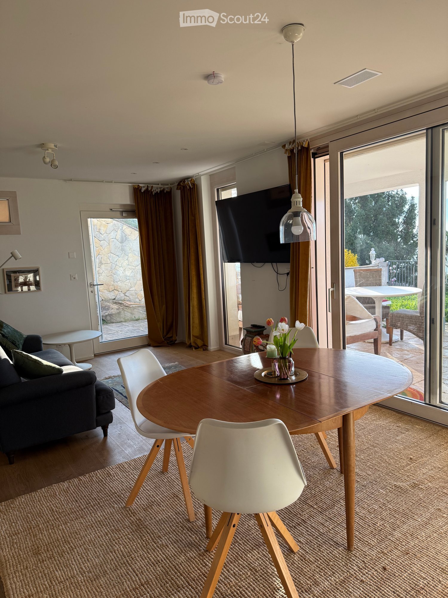Modern living and dining area with a sofa, dining table, chairs, and a television. Large sliding glass doors lead to a balcony with outdoor furniture.
