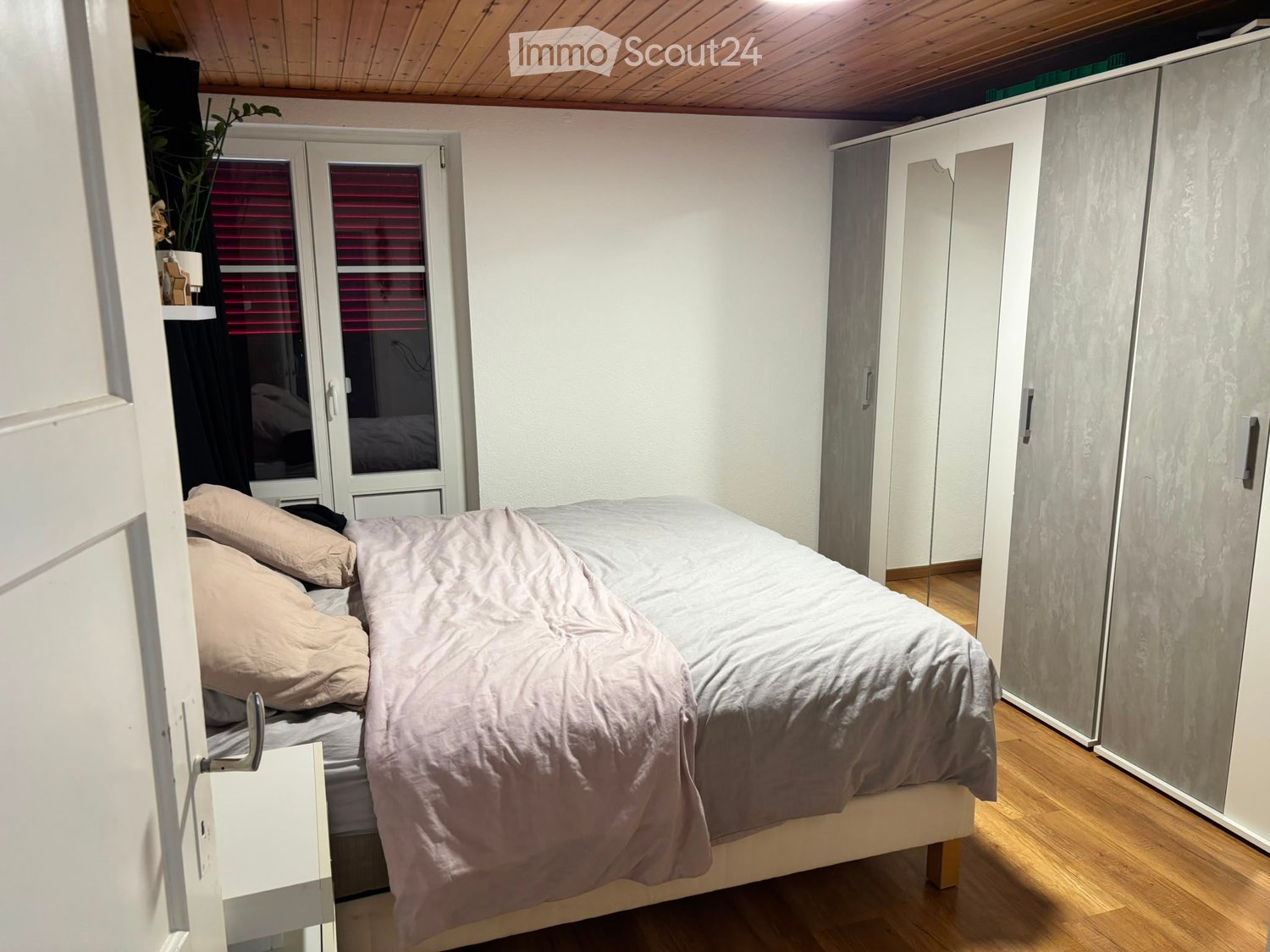 Cozy bedroom with a single bed, light-colored bedding, a large mirrored wardrobe, and a small plant on a shelf near the window.