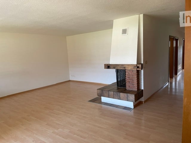 Empty room with wooden flooring, white walls, and a fireplace with a brick surround.