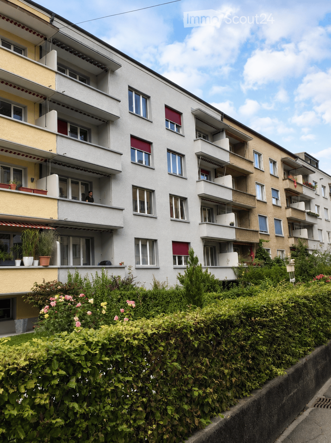Multi-story residential building with balconies, large windows, and surrounding greenery.