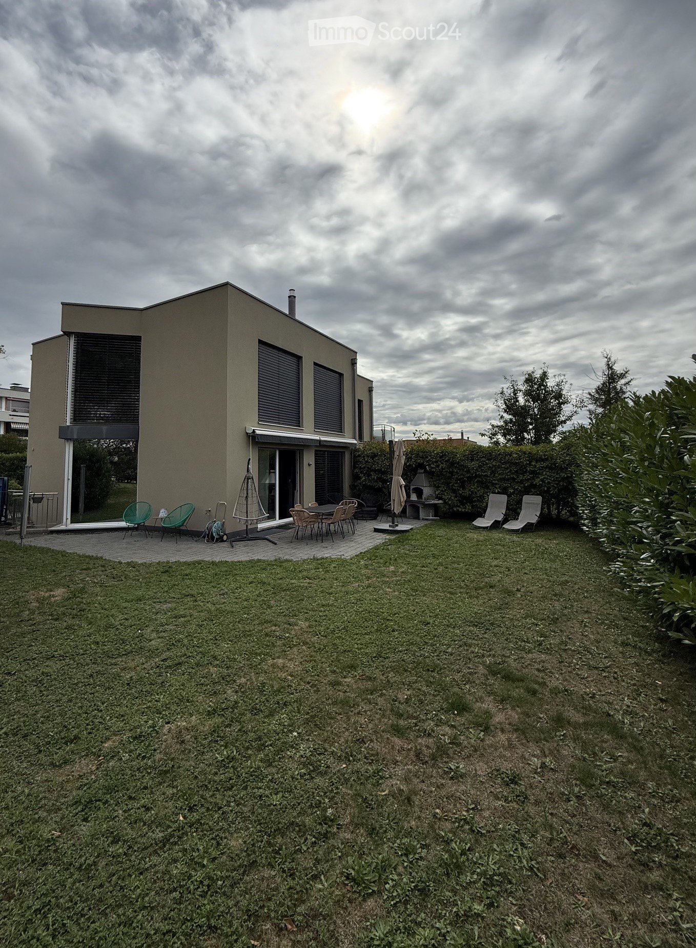 A modern house with a spacious garden area featuring a patio with chairs and a table, surrounded by greenery and a lawn.