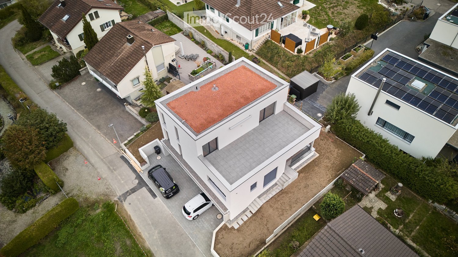 Aerial view of a modern white house with a flat roof, garage, and parking area. The house has large windows and a contemporary design. Solar panels are visible on neighboring roofs.