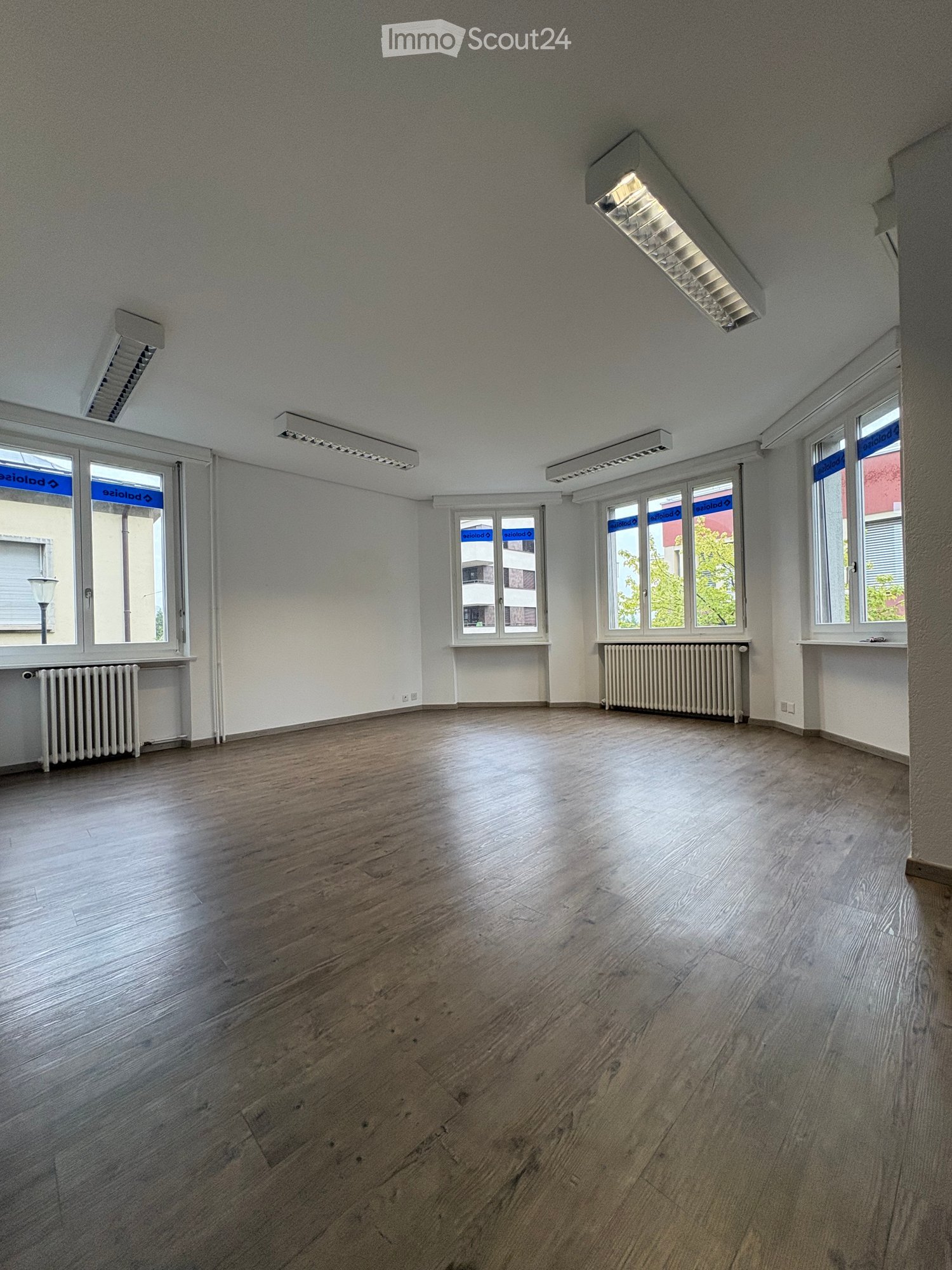 Empty room with wooden flooring, white walls, large windows, radiators, and ceiling lights.
