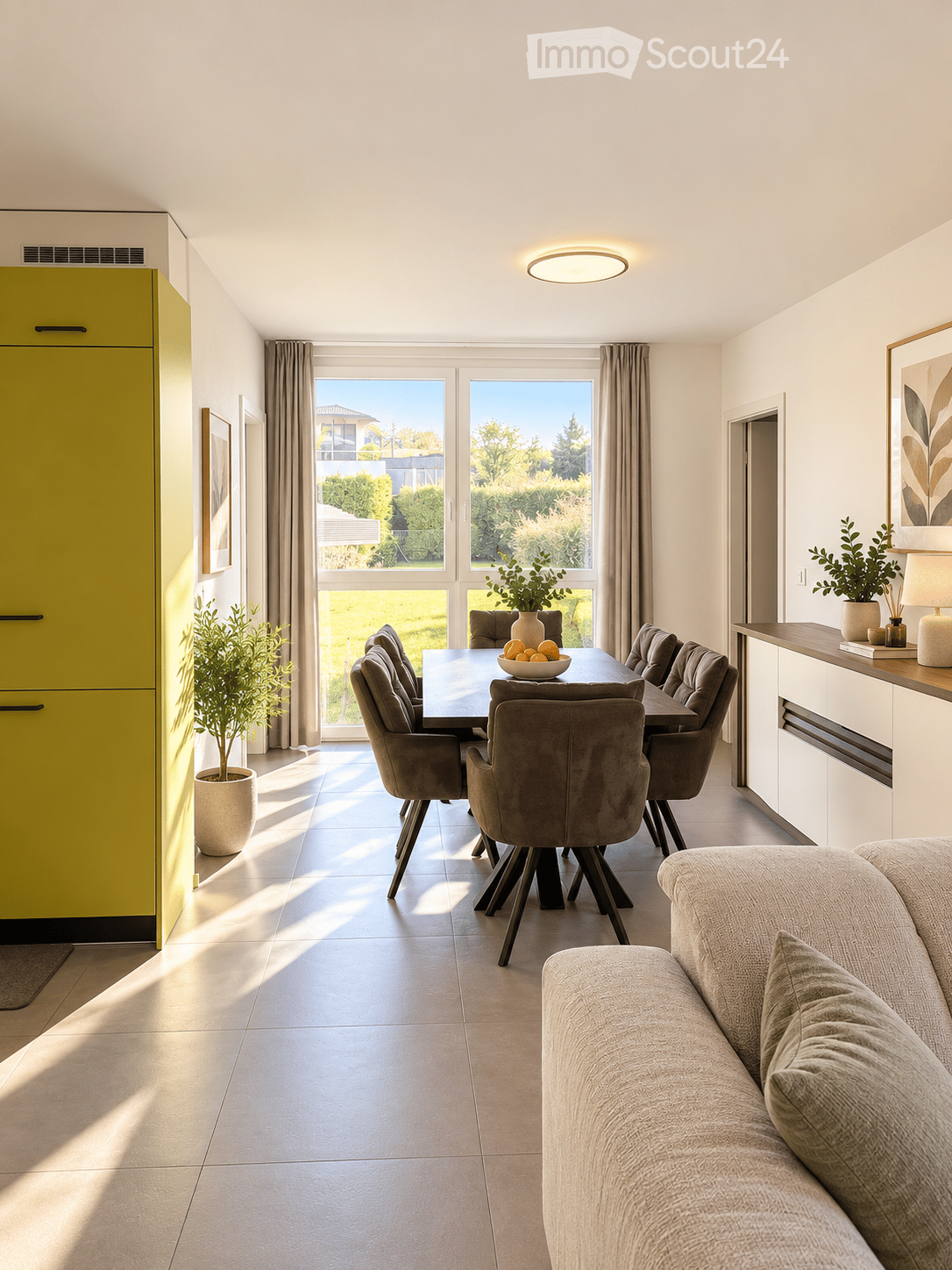 Bright, modern living and dining area with large windows, a sofa, dining table with chairs, potted plants, and a bowl of fruit on the table.