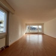 Completely empty room with wooden flooring, white walls, and large windows with a view of the city.