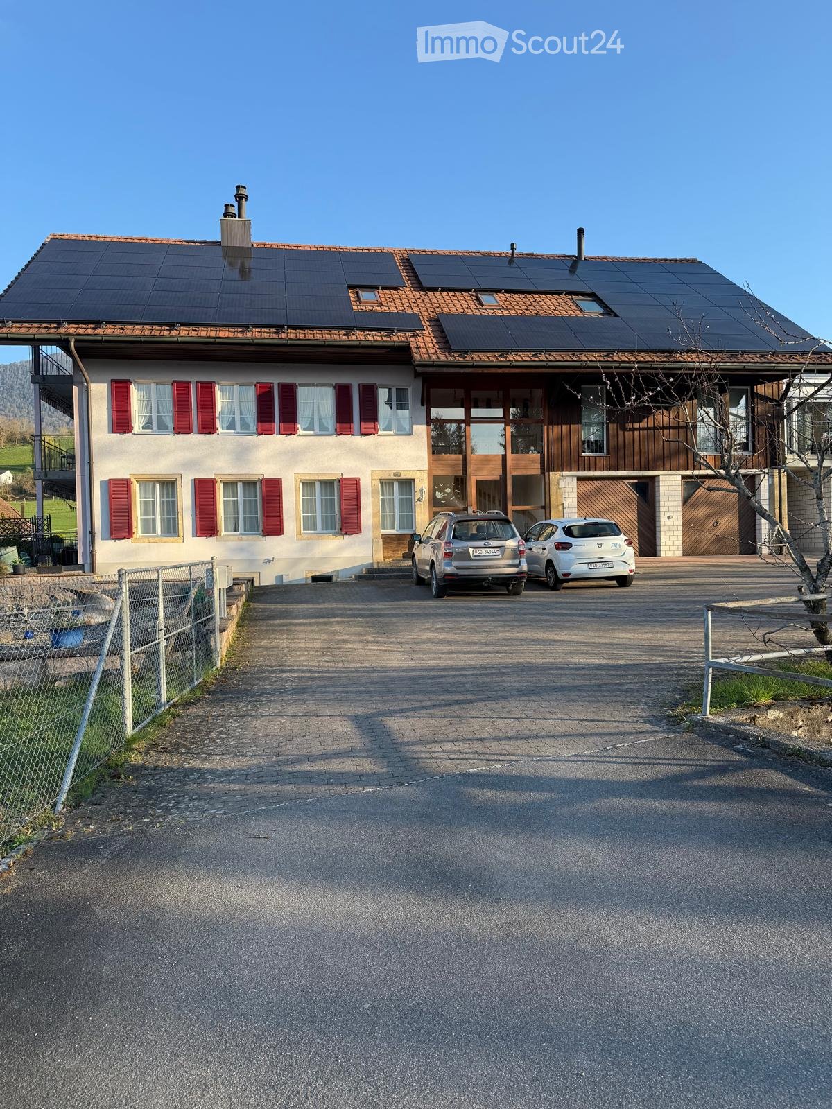 Two-story house with red shutters, solar panels on the roof, and a driveway with two parked cars.