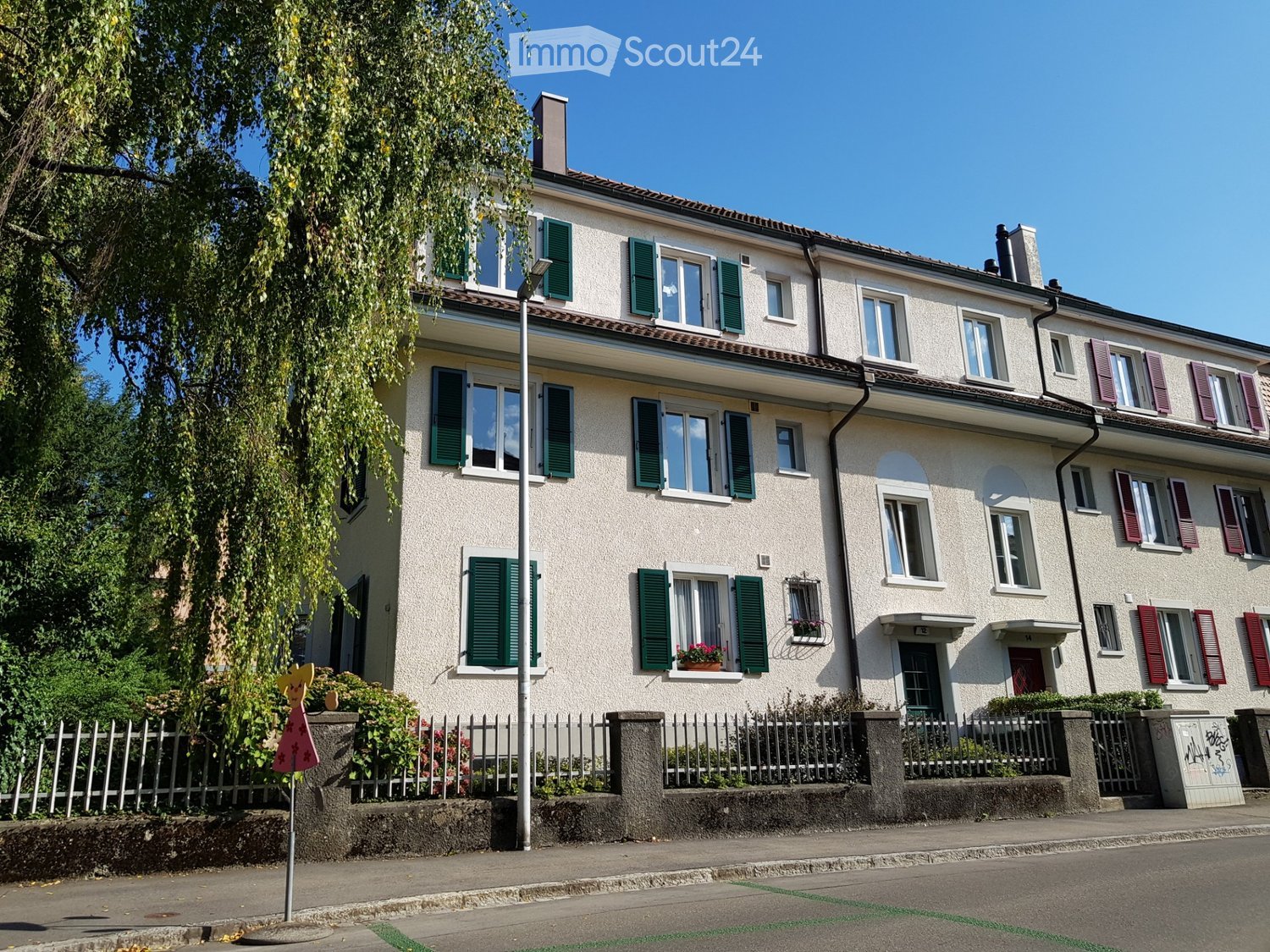 Two-story residential house with white facade, green shutters, and multiple windows. Features a small garden area with a fence and some plants.