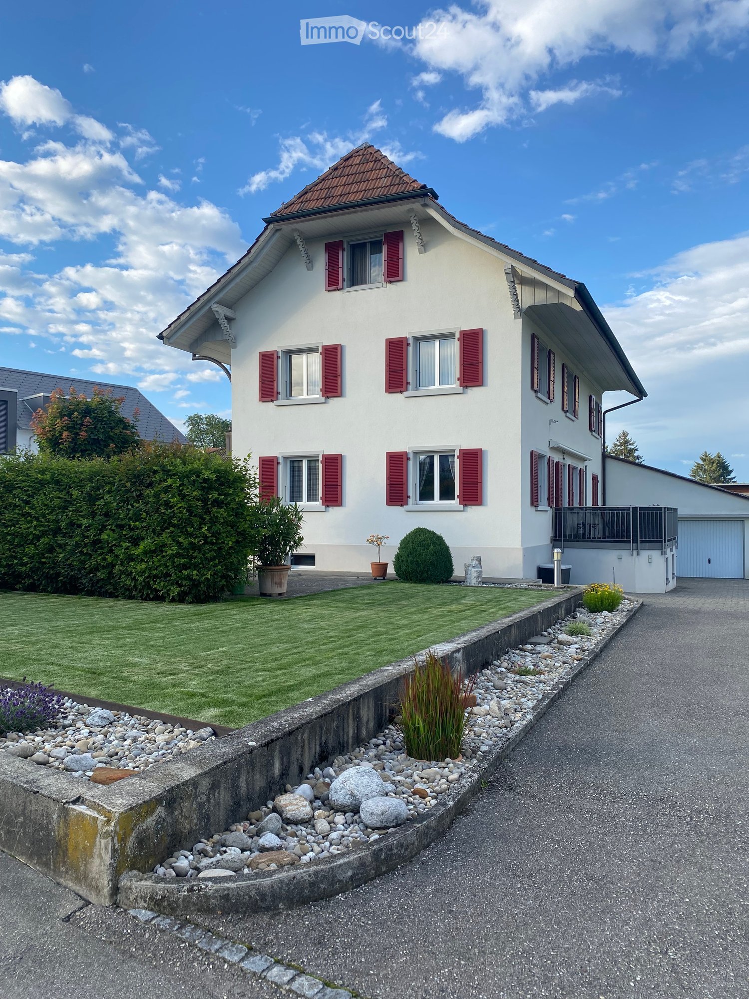 Two-story house with white walls, red shutters, and a well-maintained garden with a gravel pathway.
