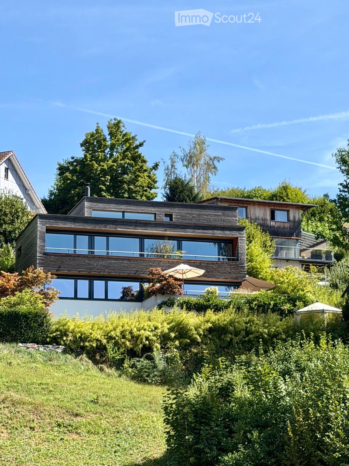 Modern two-story house with large windows and wooden exterior, surrounded by lush greenery and trees.