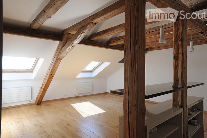 Bright, empty attic room with wooden beams and exposed wooden ceiling. Features skylight windows, wooden flooring, and a small built-in shelf unit.