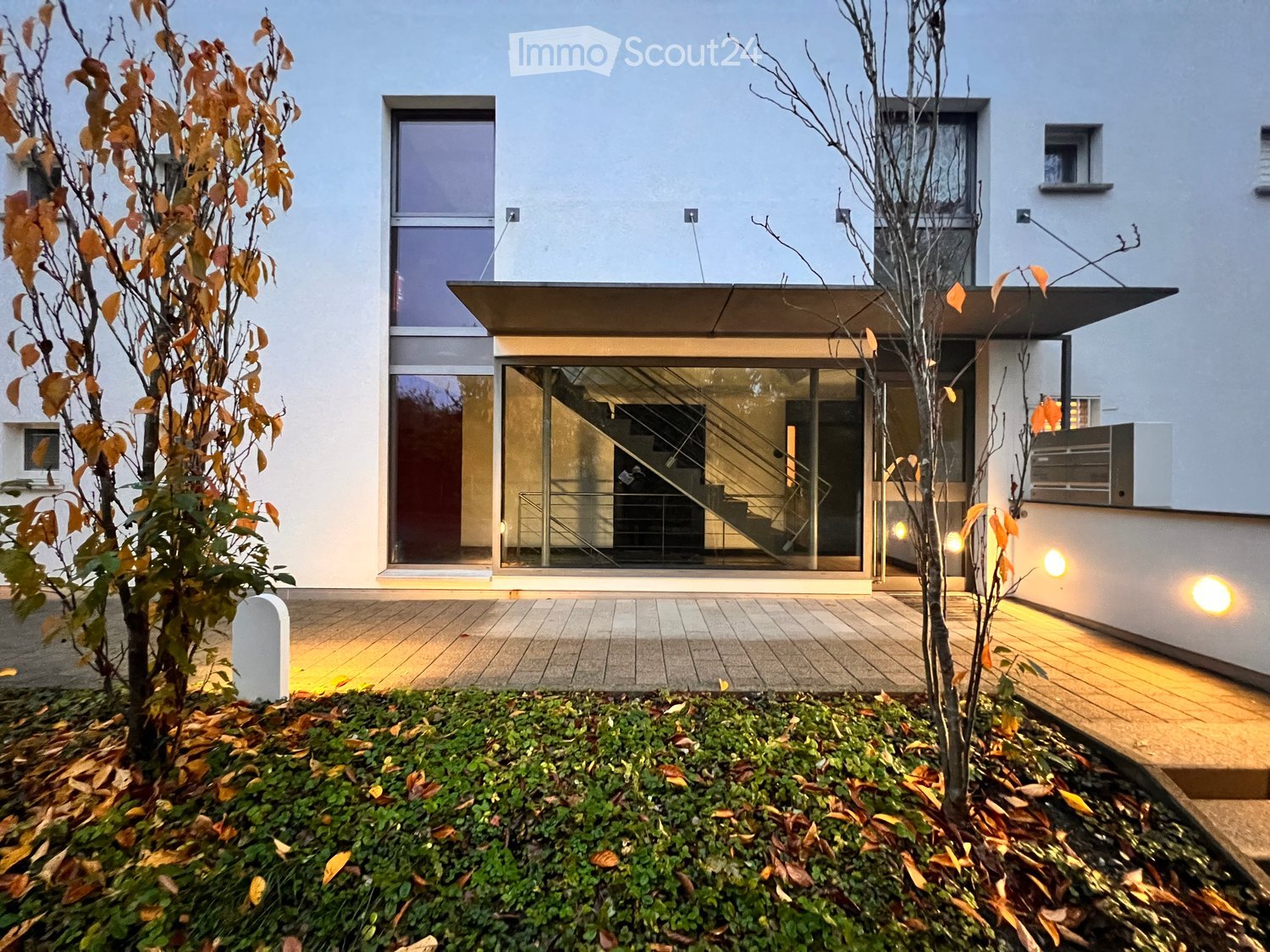 Modern two-story building with large glass windows and a raised ground floor entrance, surrounded by small trees and low shrubs.