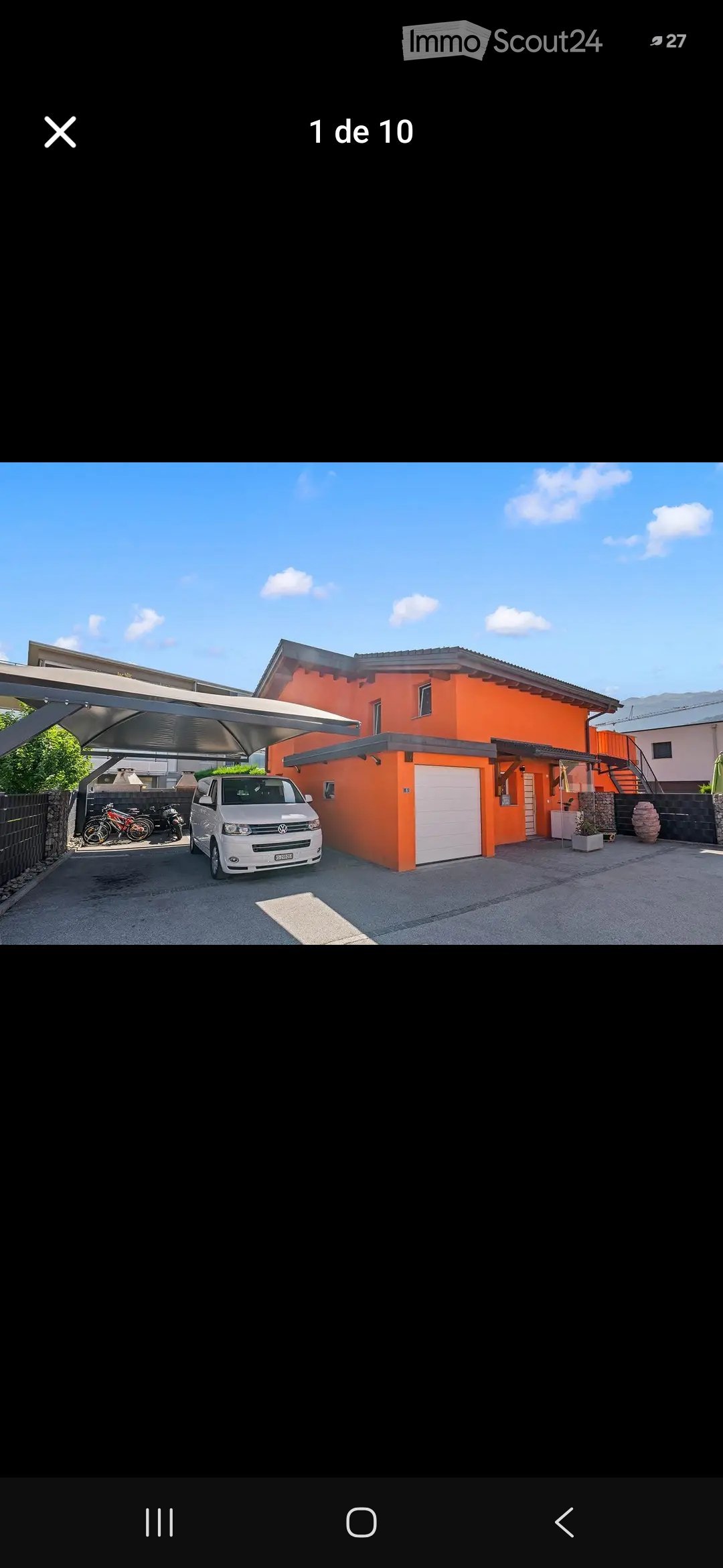 A modern two-story house with a bright orange facade, white garage door, and covered parking area. A white van is parked under the carport, and there are bicycles and motorcycles nearby.