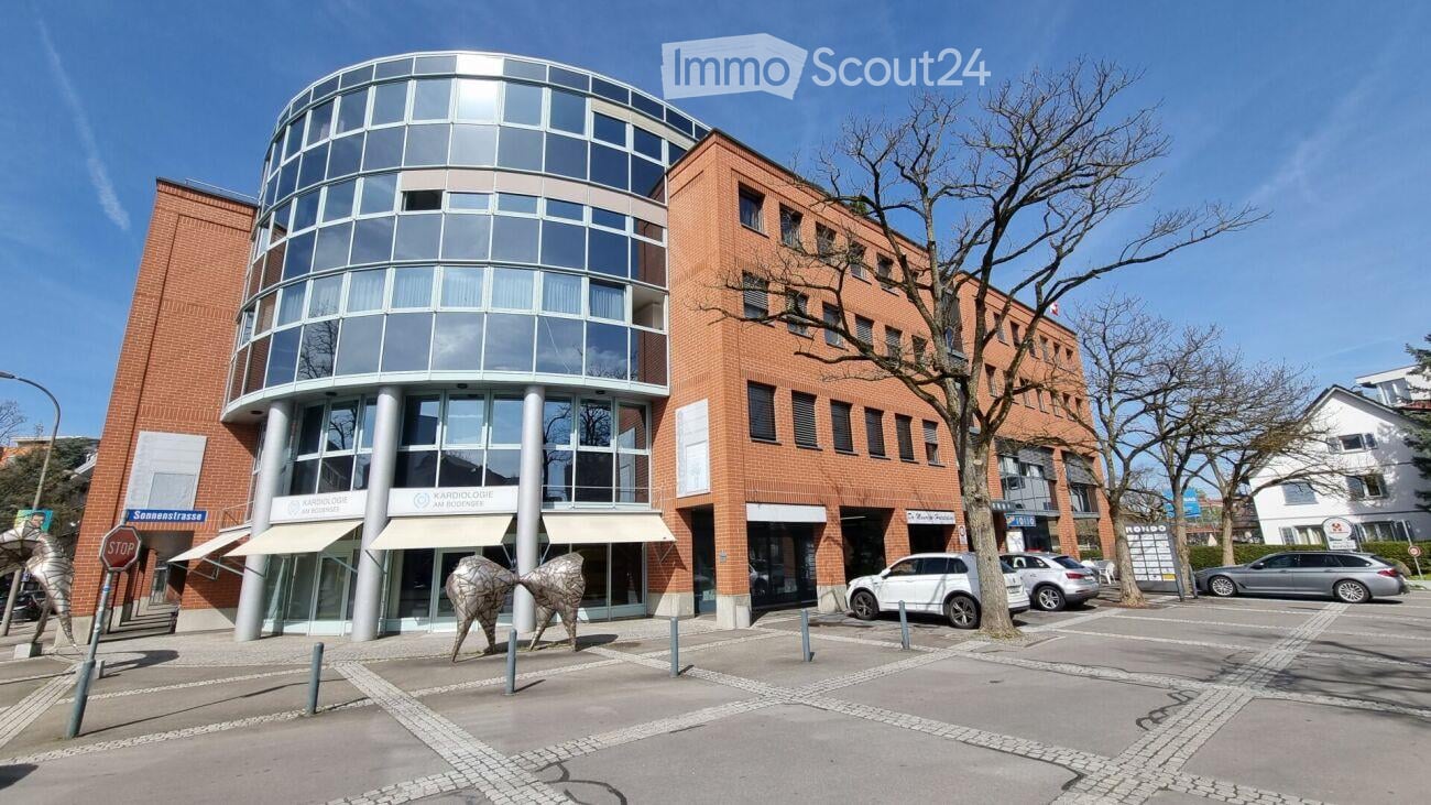 Modern multi-story building with a mix of red brick and glass facade, featuring a rounded glass tower section and several parked cars in front.