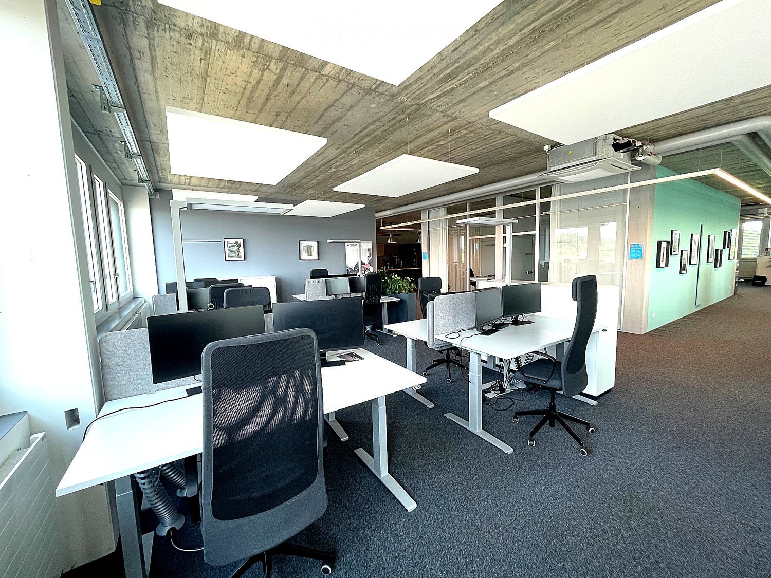 Modern office space with desks, chairs, computer monitors, and a large screen. The room features large windows, a grey carpeted floor, and a contemporary design with white and grey furniture.