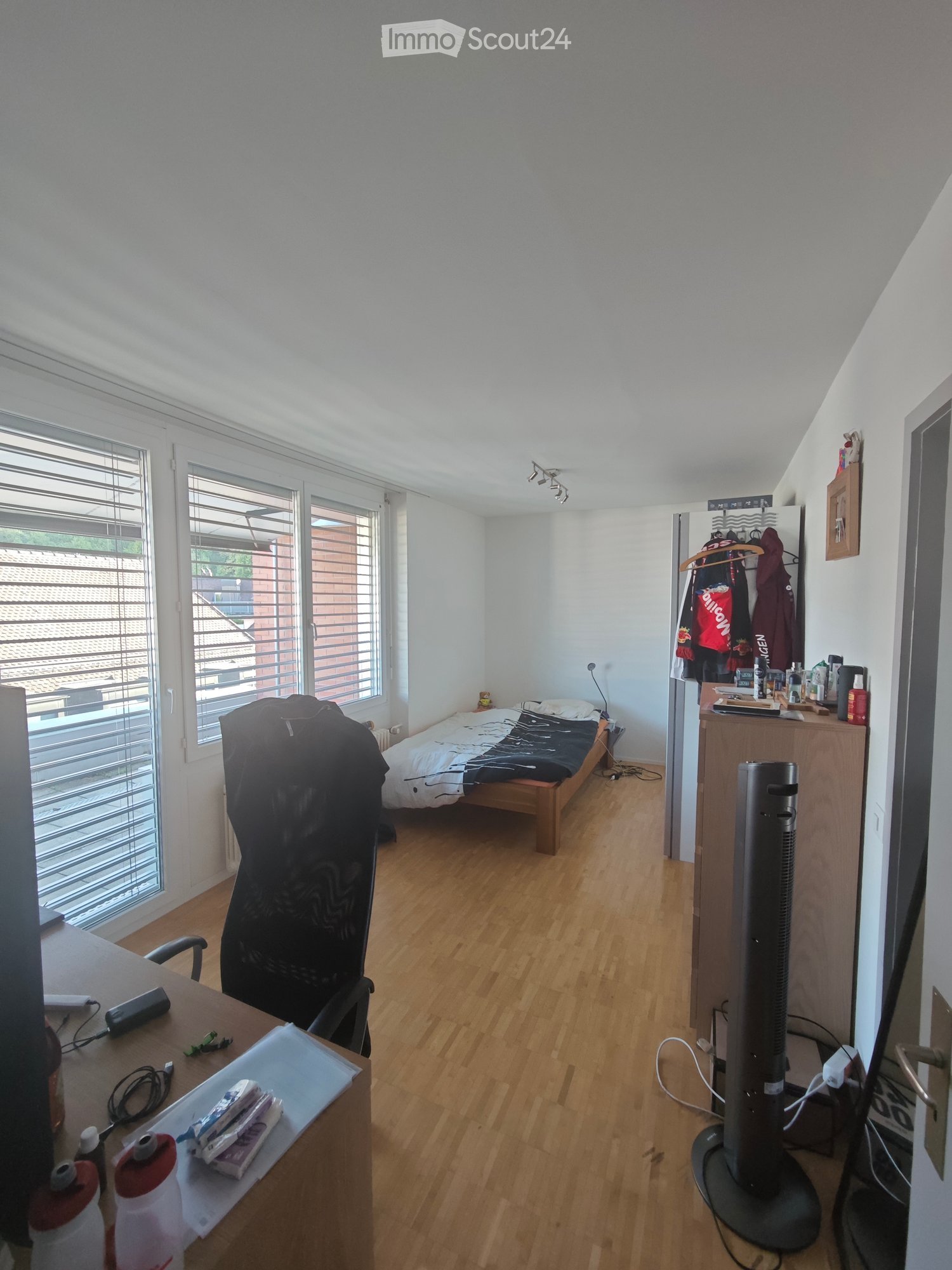 A well-lit bedroom with wooden flooring, a single bed, a desk with a chair, a fan, and a small refrigerator with a clothing rack.