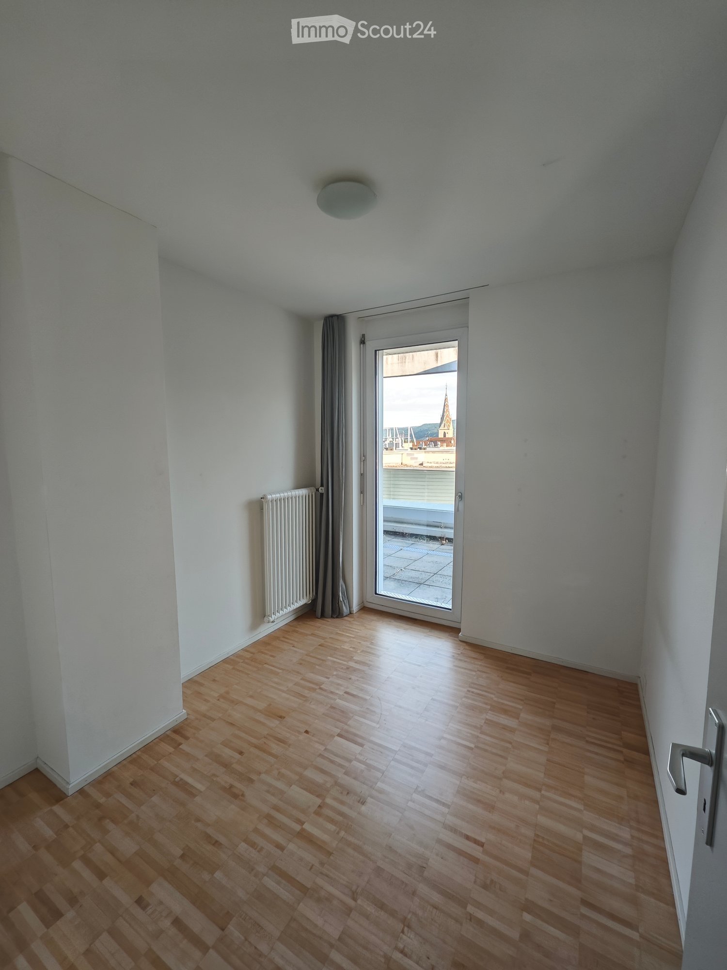 Empty room with wooden flooring, white walls, a radiator, and a sliding glass door leading to a balcony with a view of a cityscape.