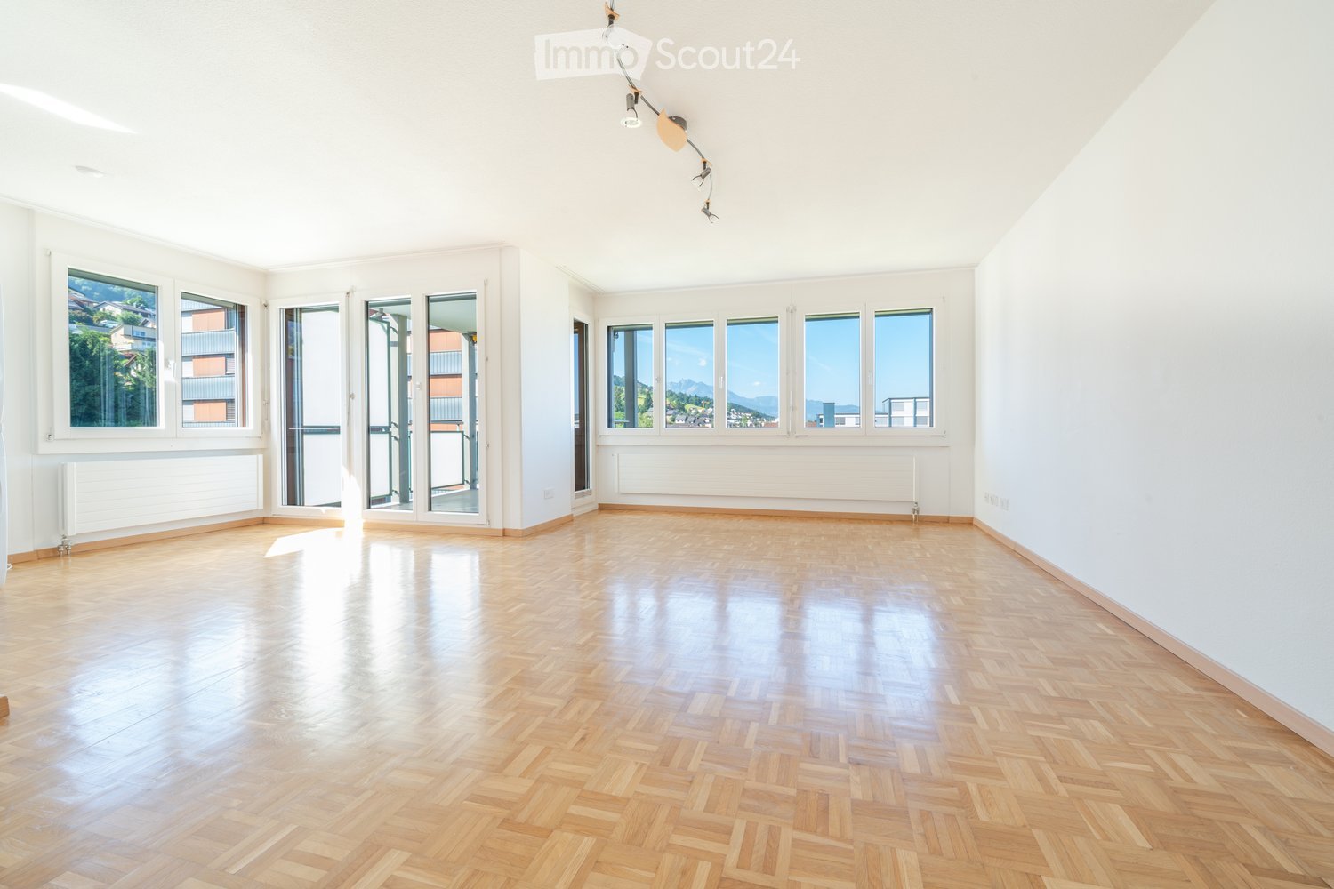 Empty room with wooden herringbone flooring, white walls, large windows, and radiators. No furniture or equipment present.