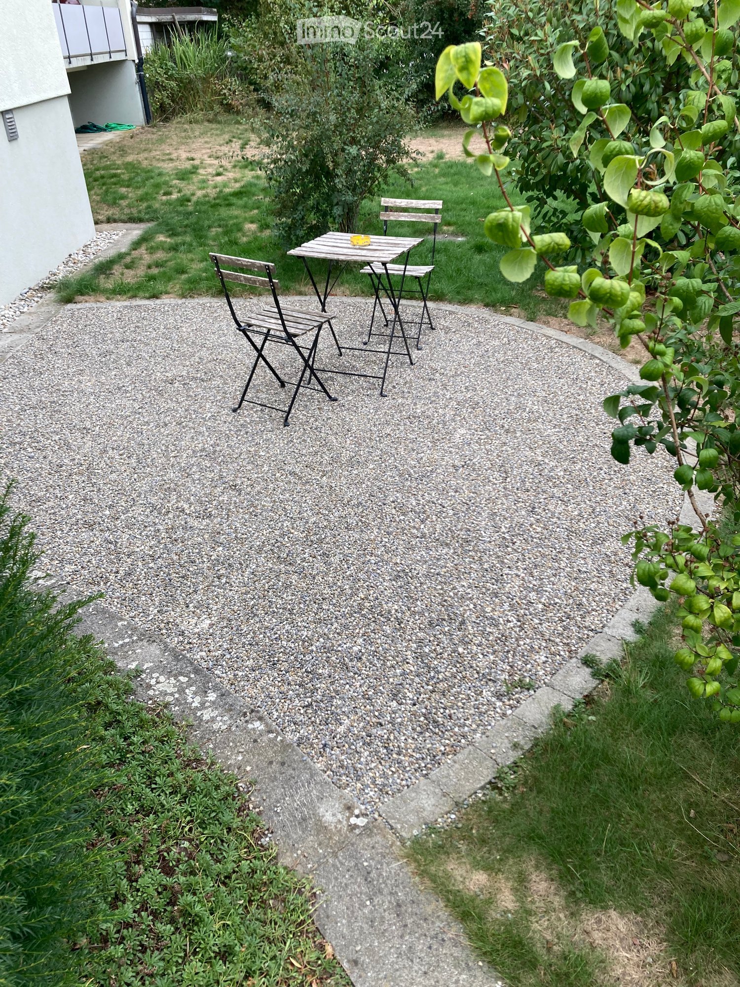 Garden area with gravel pathway, small table and chairs, surrounded by greenery and a building.