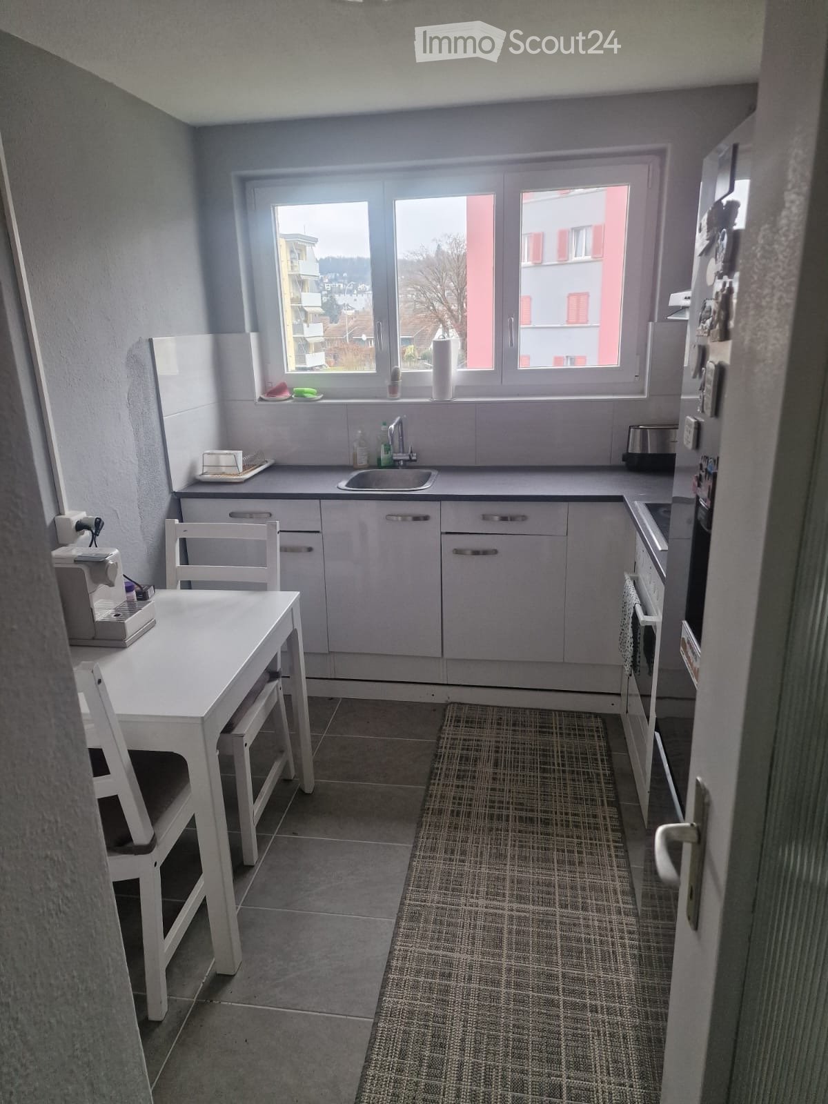Modern kitchen with white cabinets, a sink, a refrigerator, an oven, a small dining table with chairs, and a tiled floor.