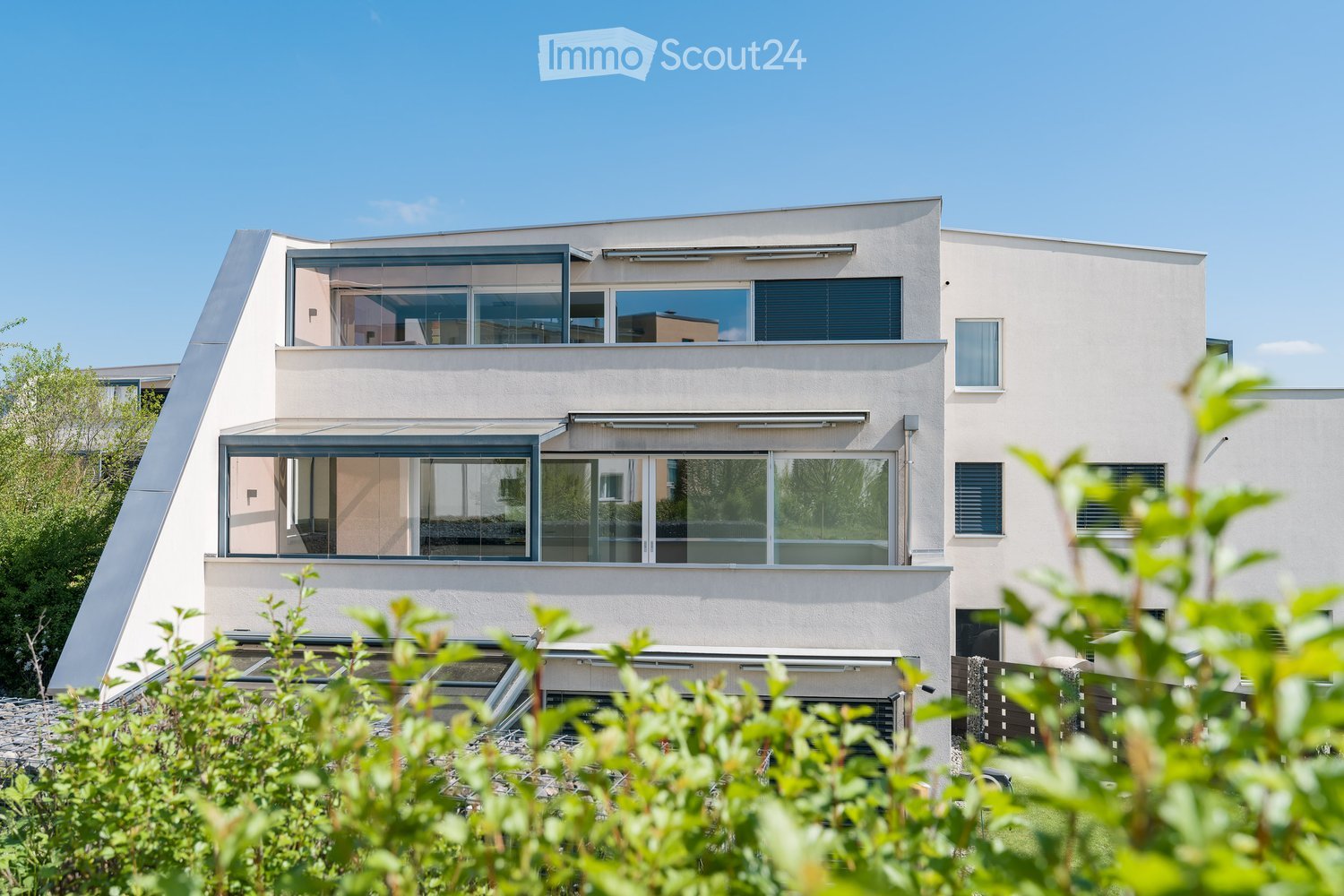Modern two-story house with large windows, clean lines, and a contemporary design. The building features a combination of light-colored walls and dark window frames, with visible greenery in the foreground.