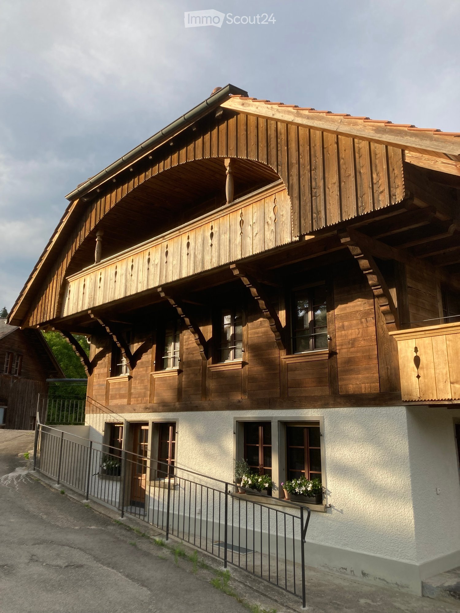 Two-story wooden house with a balcony, large windows, and decorative wooden trim. The lower level has a white plaster finish and potted plants near the entrance.