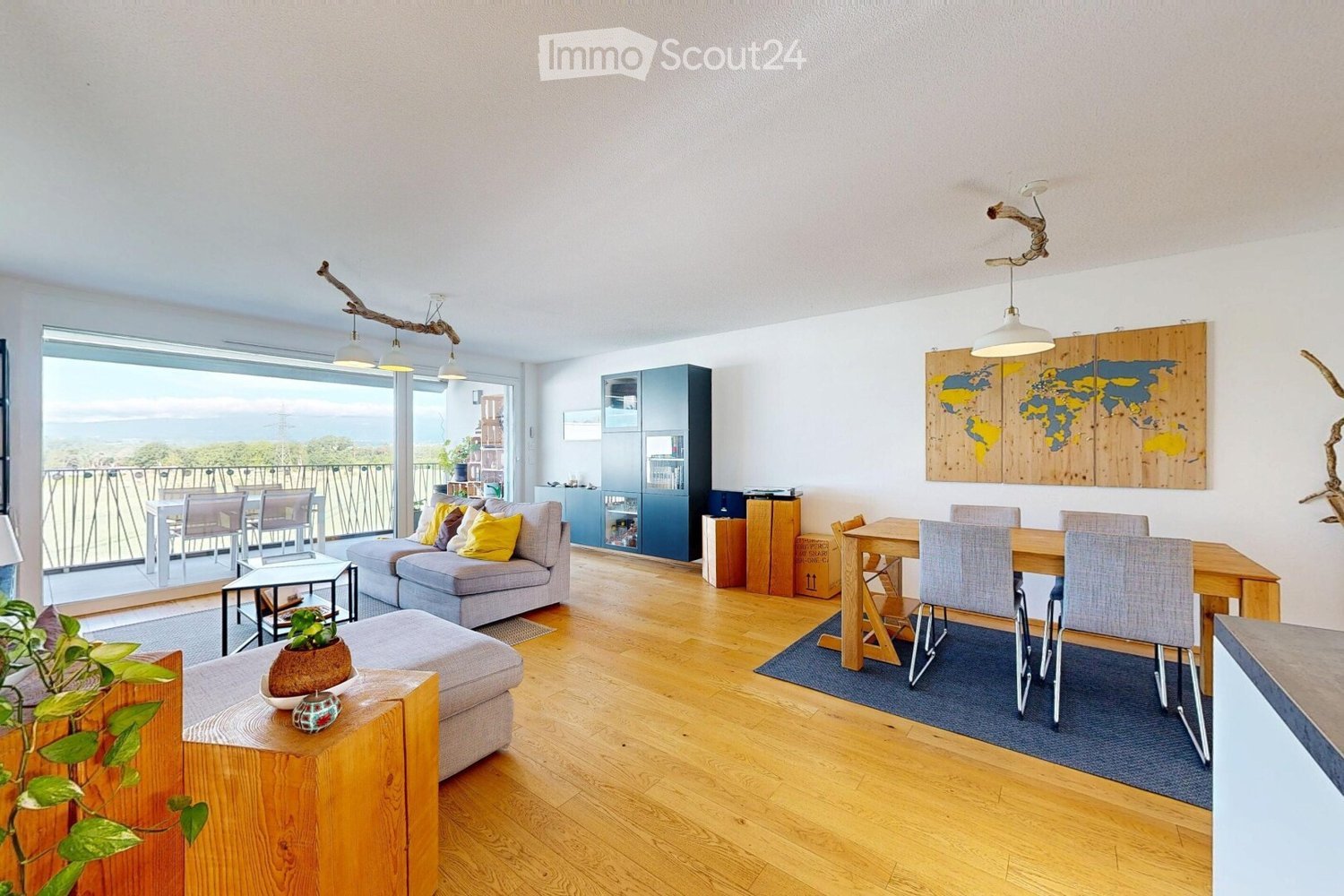 Bright living and dining area with wooden flooring, large windows offering a scenic view, a grey sofa with colorful cushions, a wooden coffee table, a dining table with chairs, and a cabinet.
