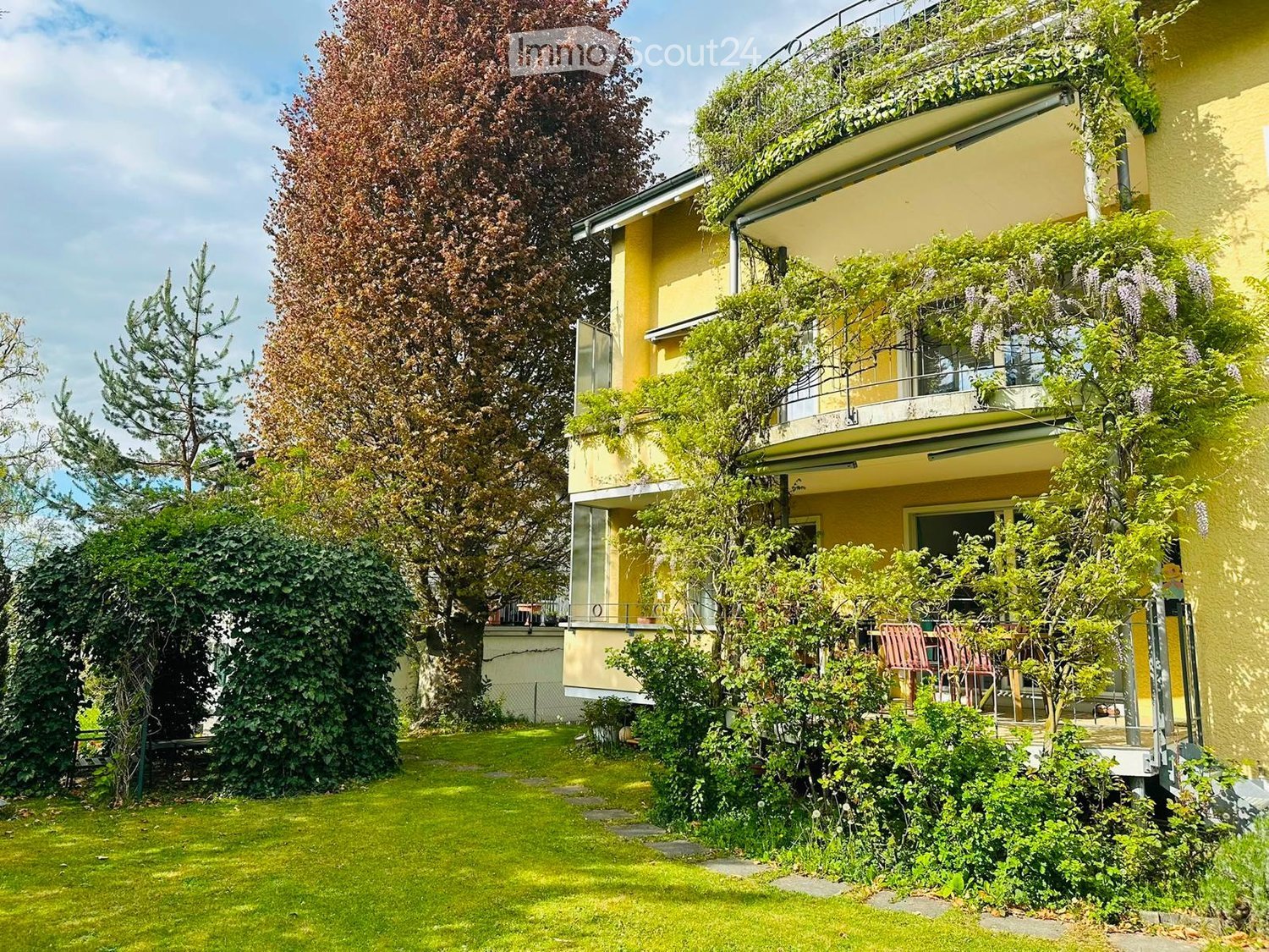 A well-maintained garden with a lawn, trees, and a multi-story yellow building with balconies and greenery on the facade.