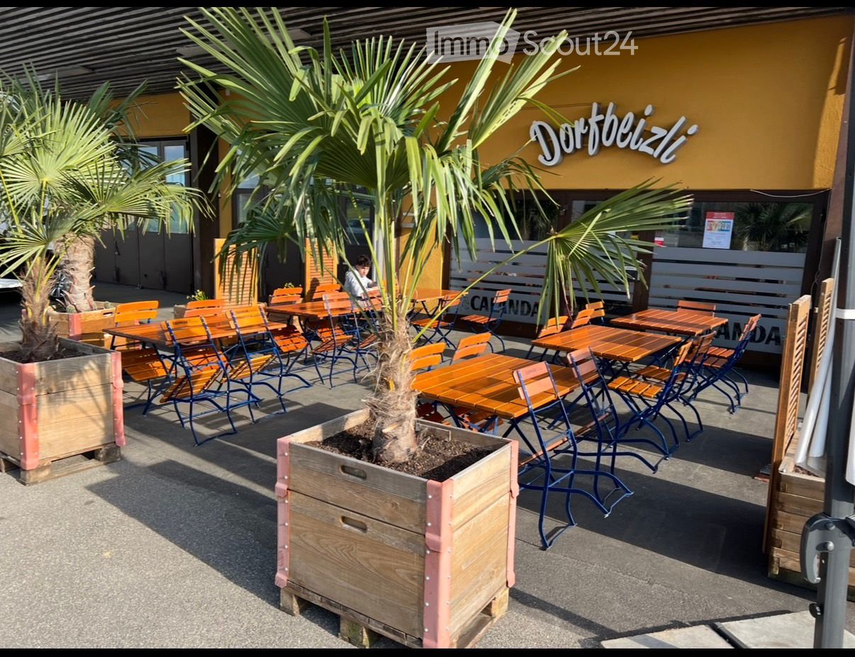 Outdoor seating area with wooden tables and chairs arranged in rows, featuring multiple potted palm trees on the ground, yellow building, signage with the text 'Dortbeizl', and an outdoor seating arrangement for customers.