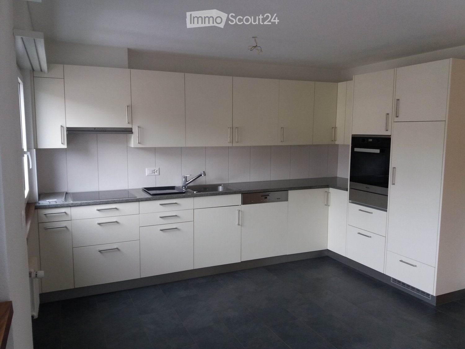 Modern kitchen with white cabinets, stainless steel sink, faucet, oven, and dishwasher. Dark tiled floor and countertops.