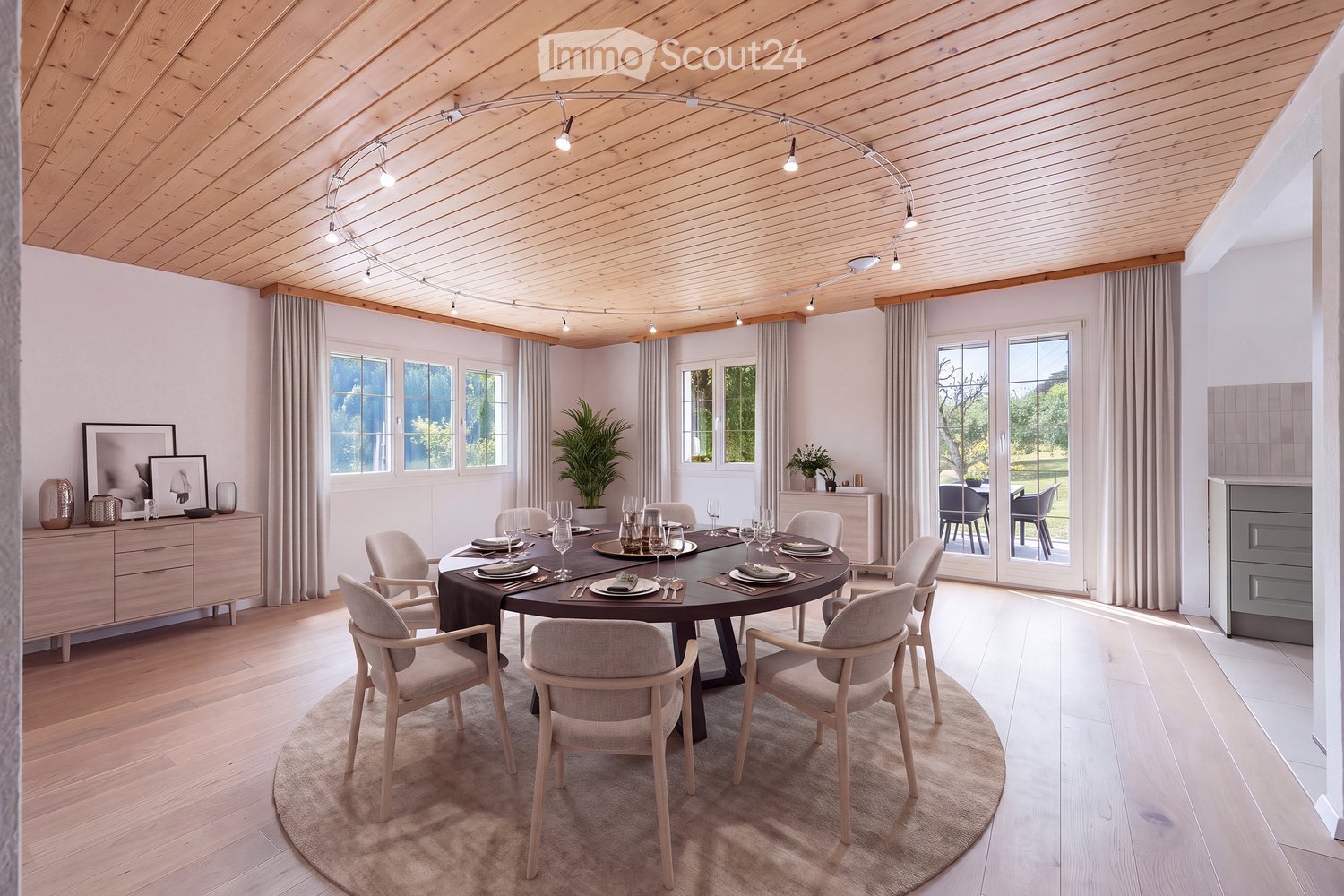 A modern dining room with a round wooden dining table surrounded by six chairs, set with plates, cutlery, and glasses. The room features light wooden flooring, white walls, large windows with curtains, and a sleek cabinet with decorative items. A potted p