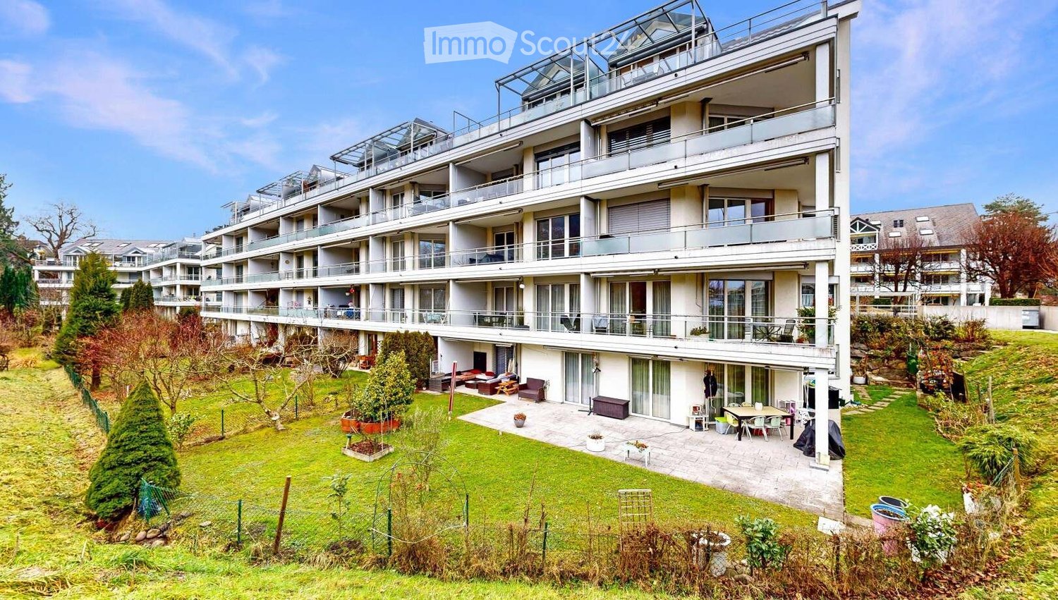 Modern multi-story residential building with balconies and a landscaped garden area featuring seating and greenery.