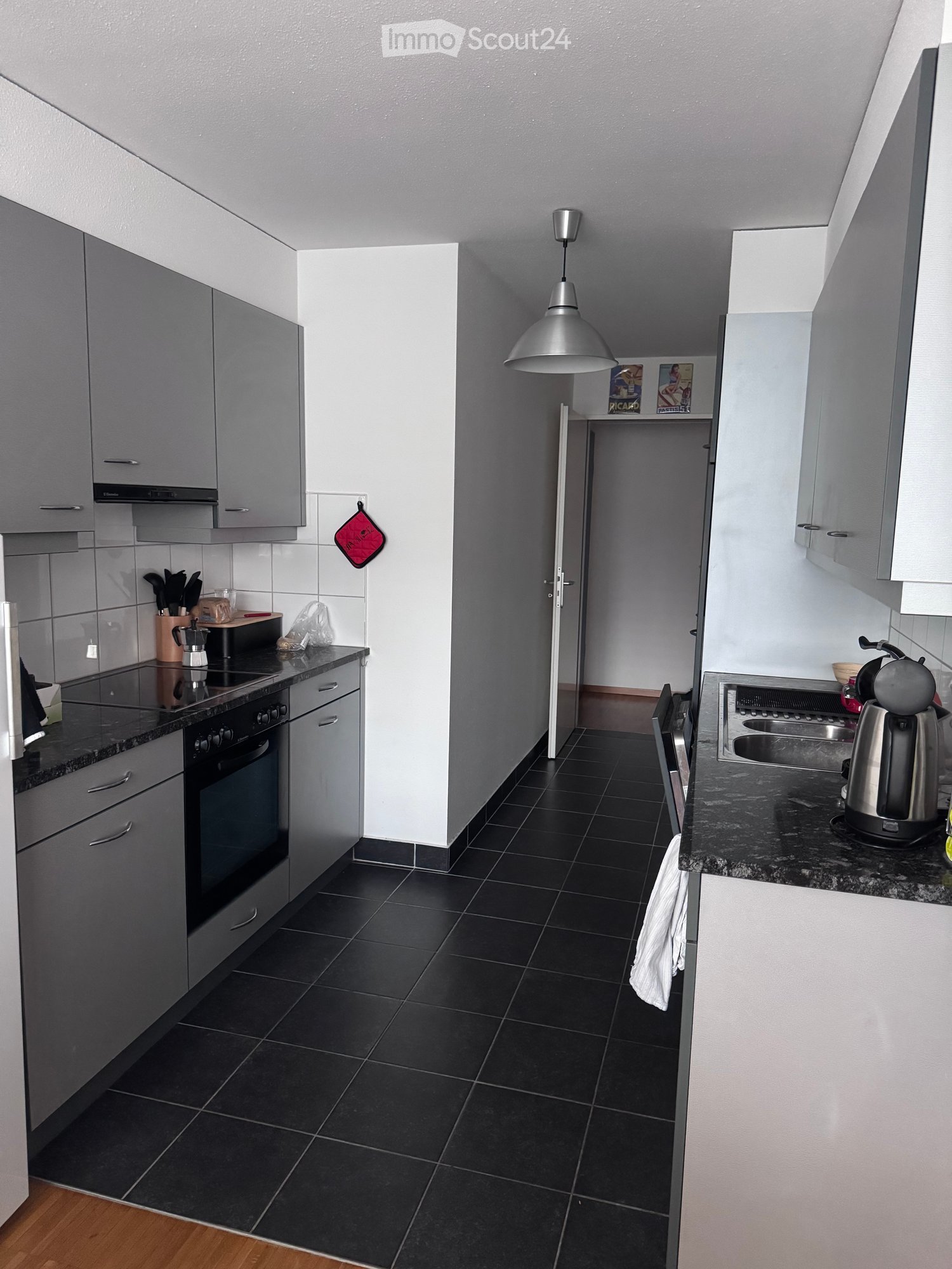 Modern kitchen with grey cabinets, black tiled floor, and stainless steel appliances including an oven, stove, and fridge. A kitchen island with a coffee maker and utensils is visible, along with a hanging pendant light and a small shelf with decorative i