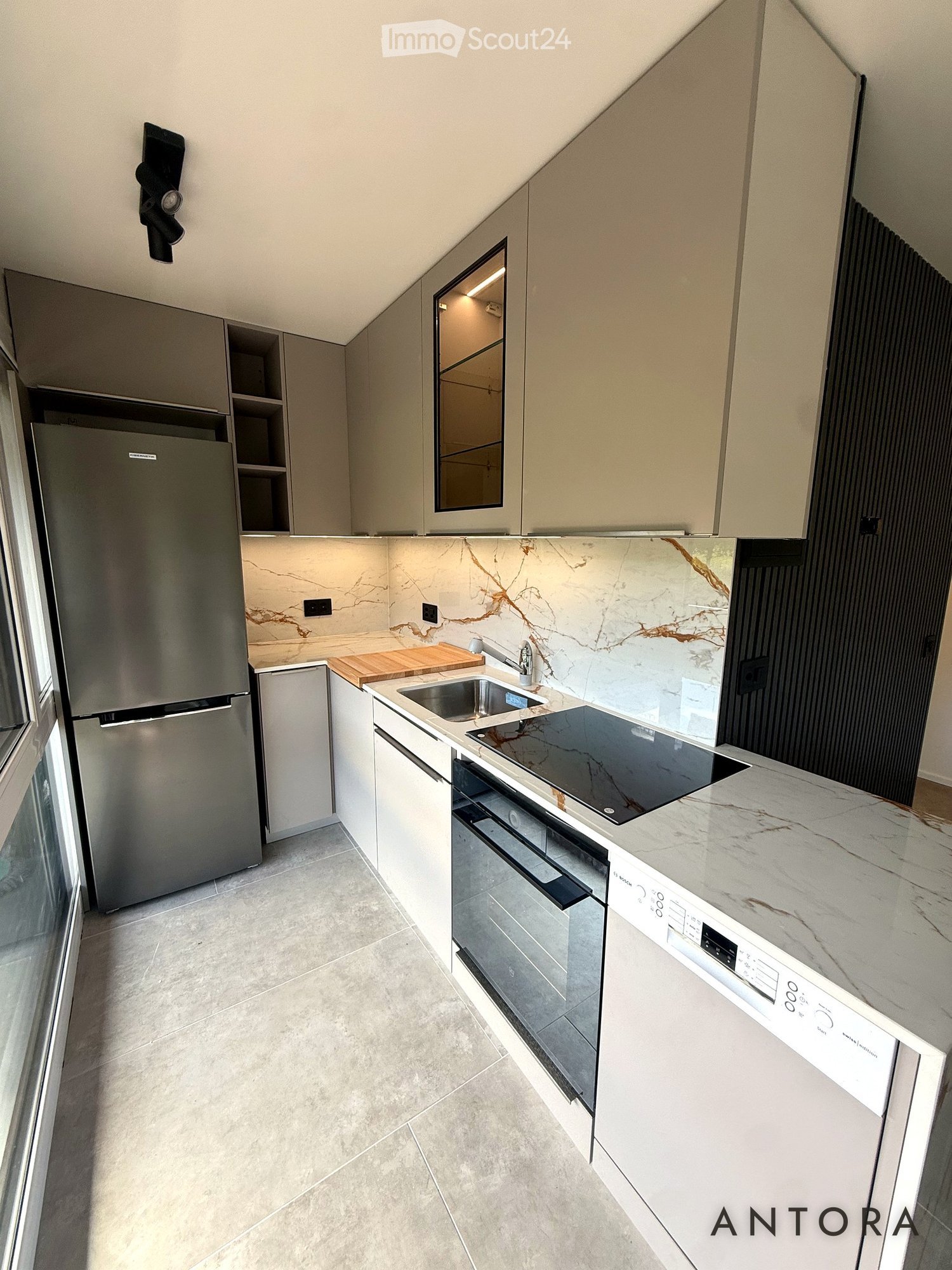 Modern kitchen with light-colored cabinets, stainless steel appliances, marble countertops, and a large window providing natural light.