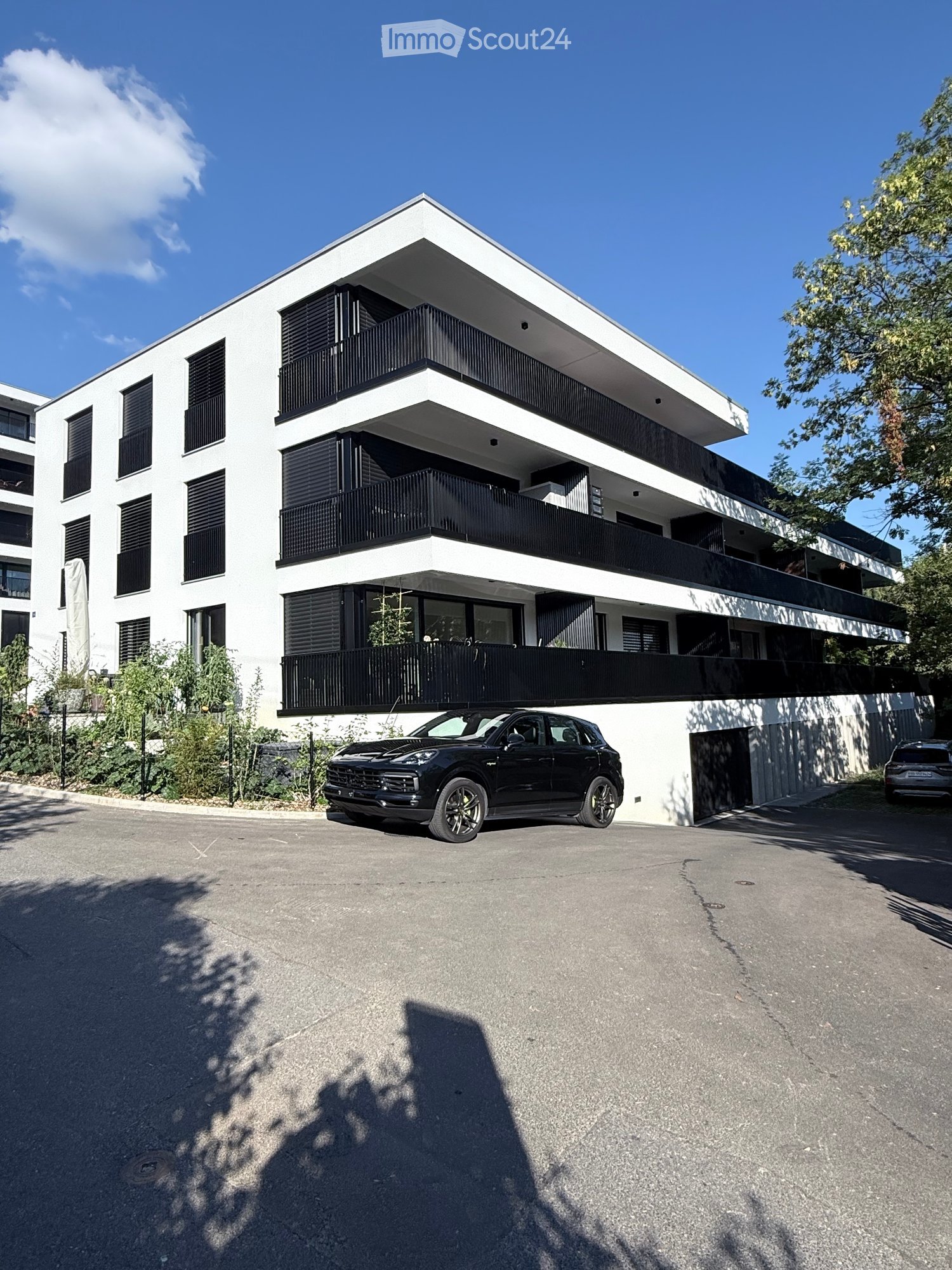 Modern three-story residential building with white facade, black balconies, and large windows. A black SUV is parked in front of the building.