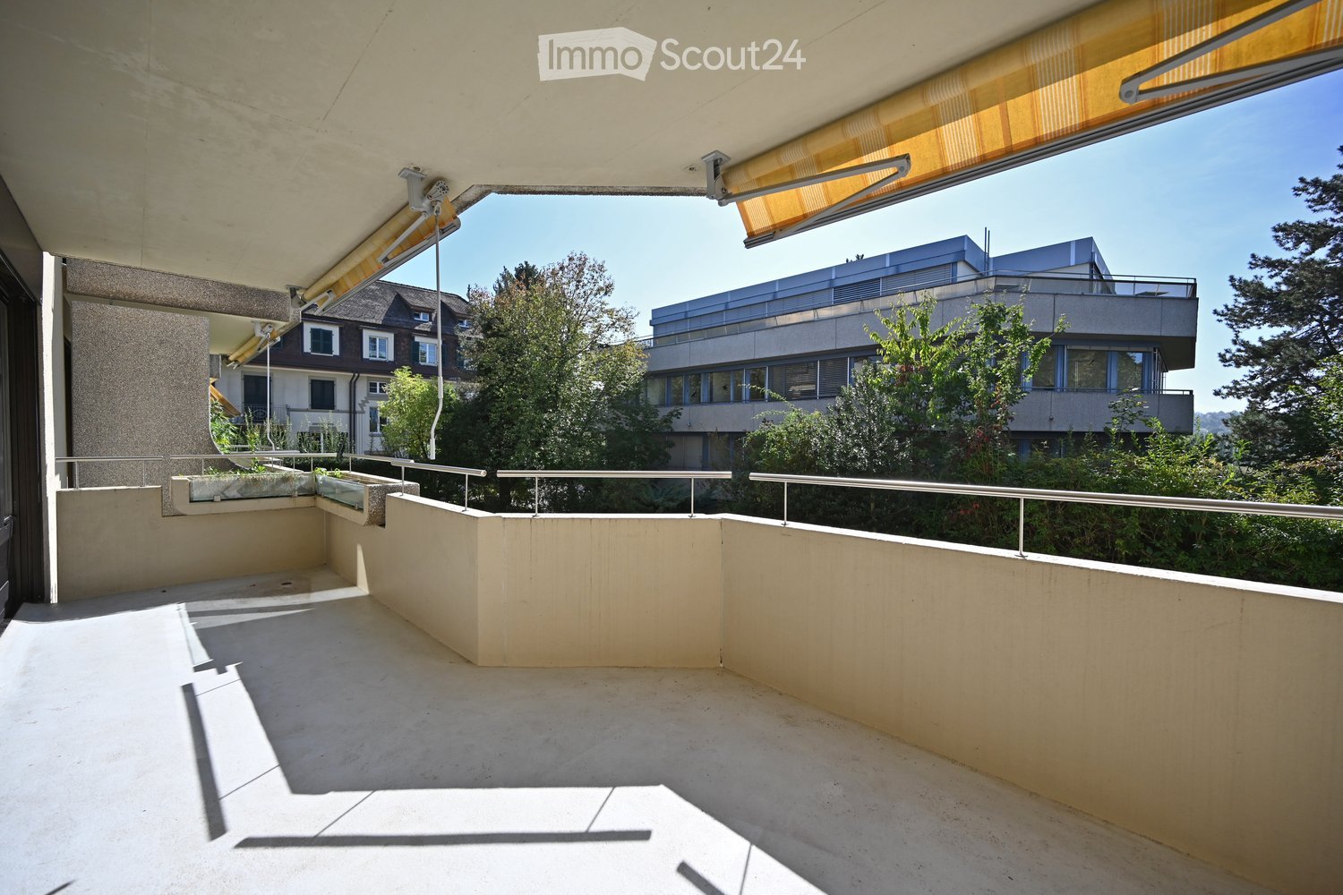 Balcony with concrete floor, metal railings, and a retractable awning, overlooking a residential area with trees and buildings.