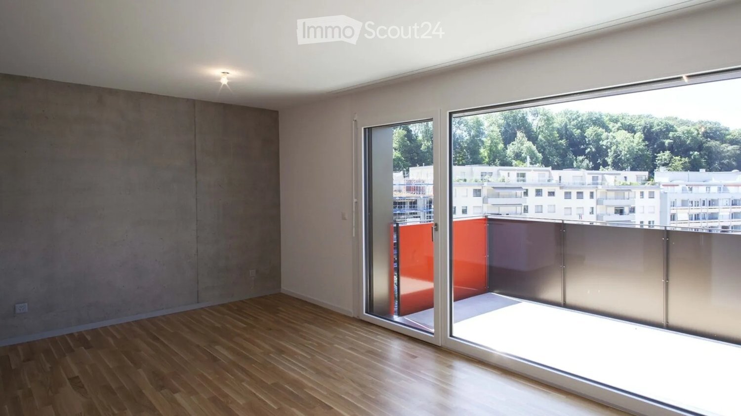 Empty room with wooden flooring, large glass sliding doors leading to a balcony, and a view of surrounding buildings and greenery.
