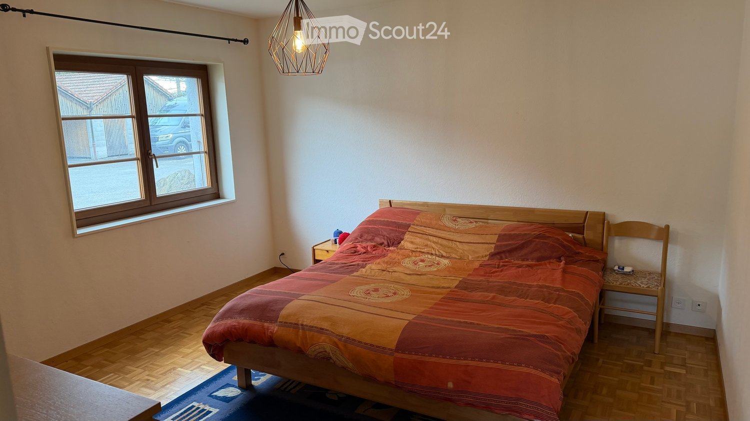 Cozy bedroom with wooden floor, large window, bed with colorful bedding, nightstand, and a wooden chair.