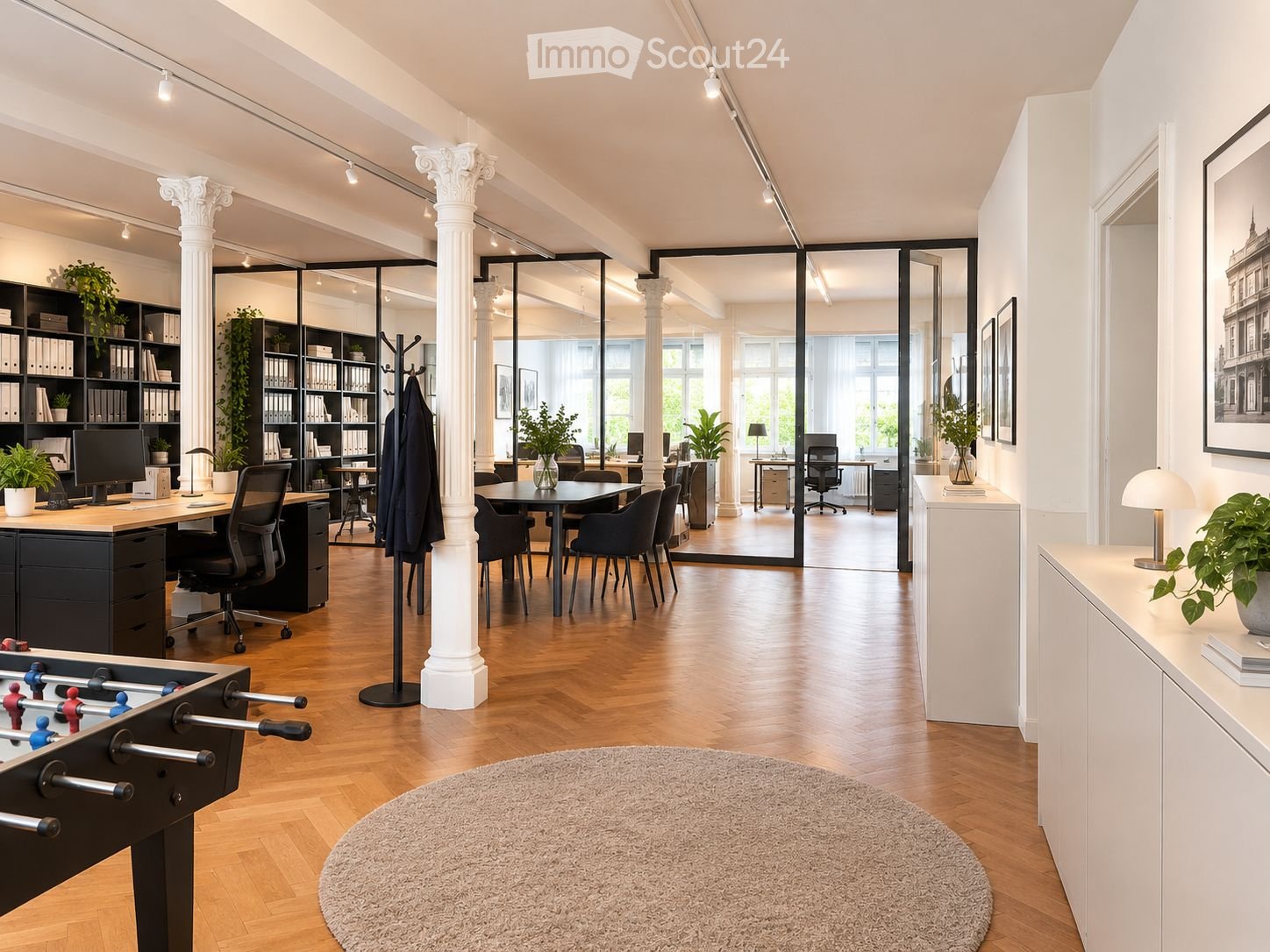 Modern office space with wooden flooring, large glass partitions, multiple desks, chairs, a football table, bookshelves, lamps, and plants.