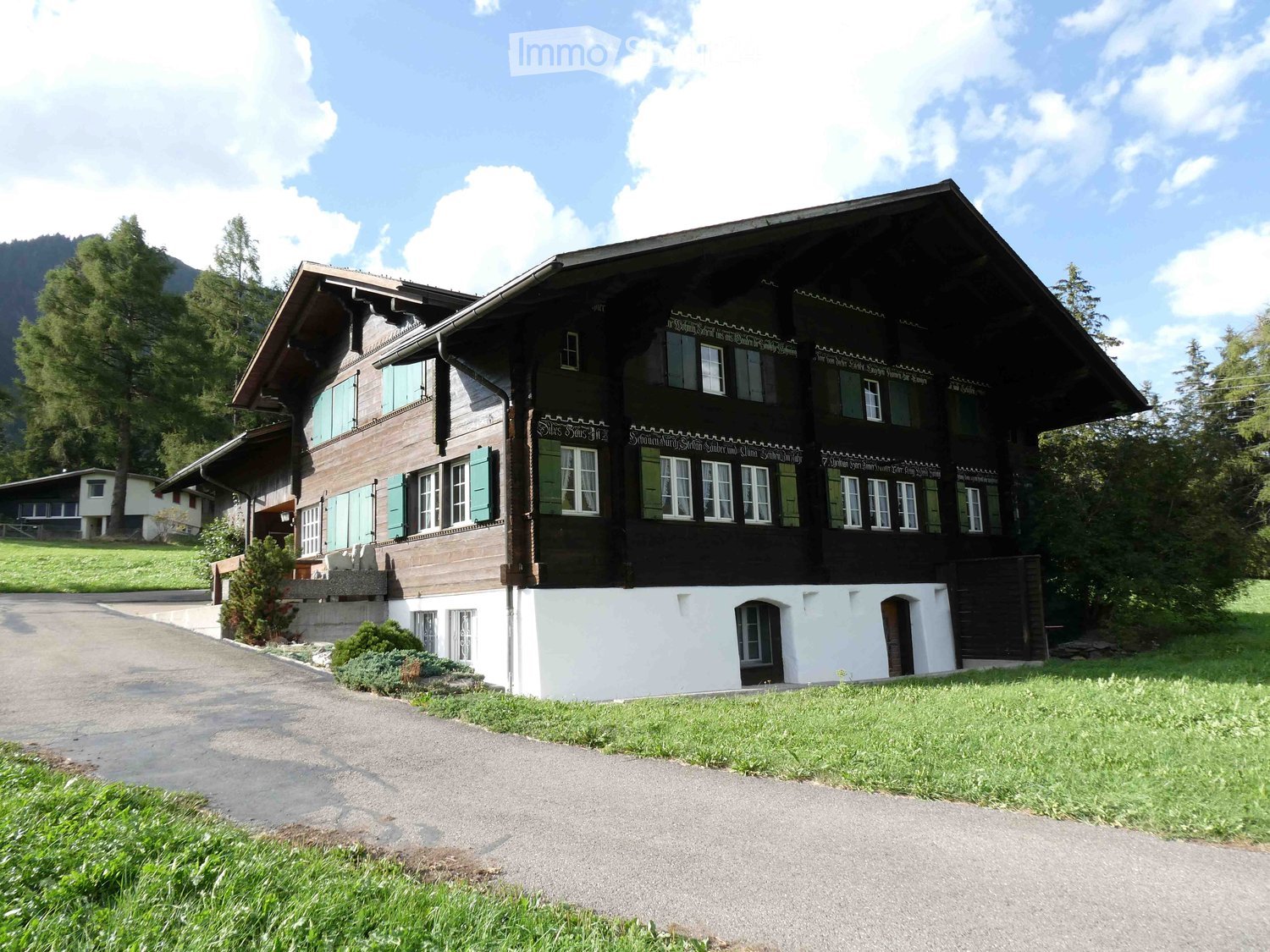 A large, traditional wooden house with a steeply pitched roof, multiple windows, and decorative wooden trim. The house is surrounded by a well-maintained lawn and trees, with a gravel driveway leading to the entrance.