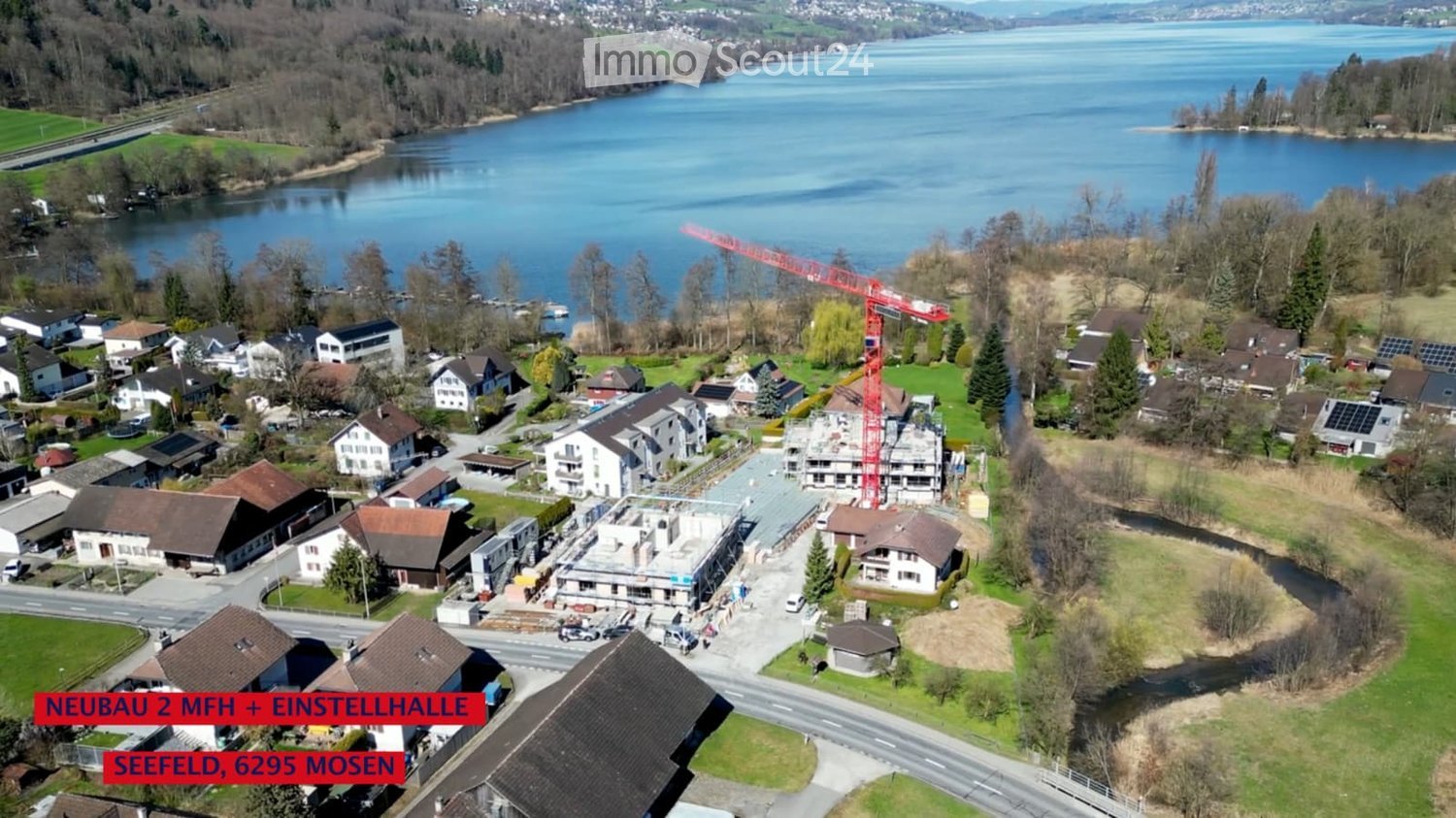 Aerial view of a residential area near a lake, featuring multiple houses, a construction site with a crane, and surrounding greenery.