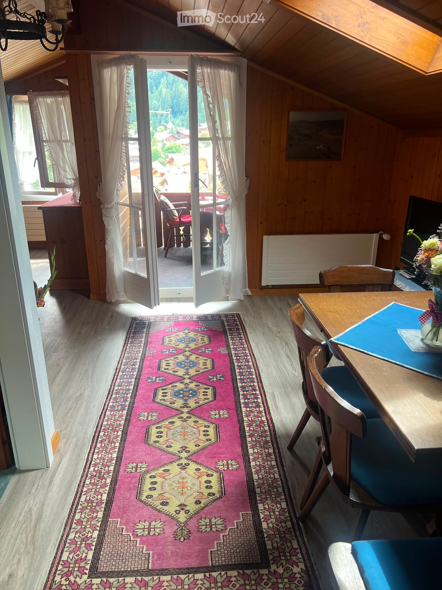 A cozy living room with wooden walls and floors, a colorful runner rug, a table with chairs, a television, and a vase with flowers. The room has large windows with curtains, leading to a balcony with outdoor furniture.